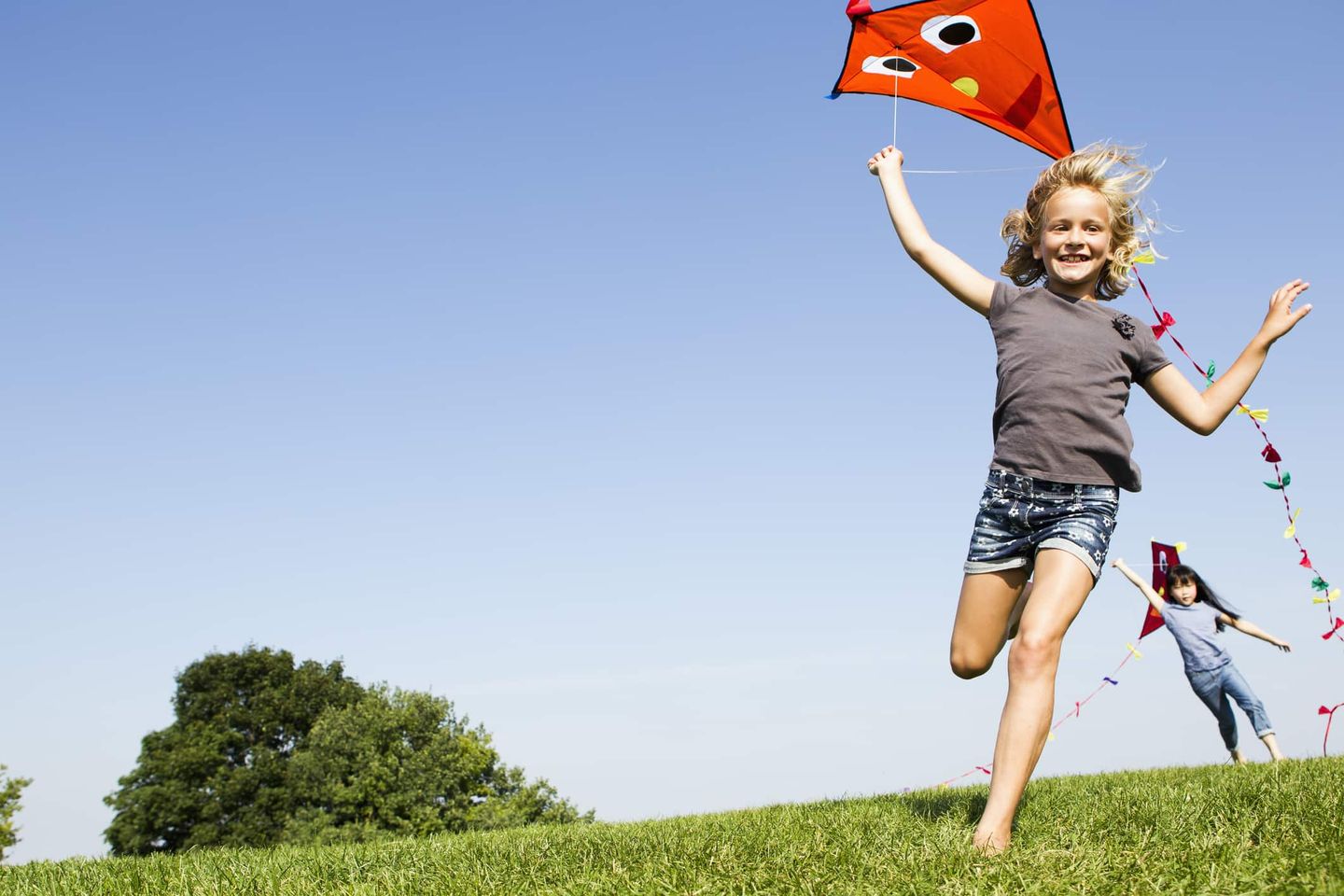 Zwei Kinder rennen lachend über eine Wiese und lassen rote Drachen mit langen Schwänzen fliegen.