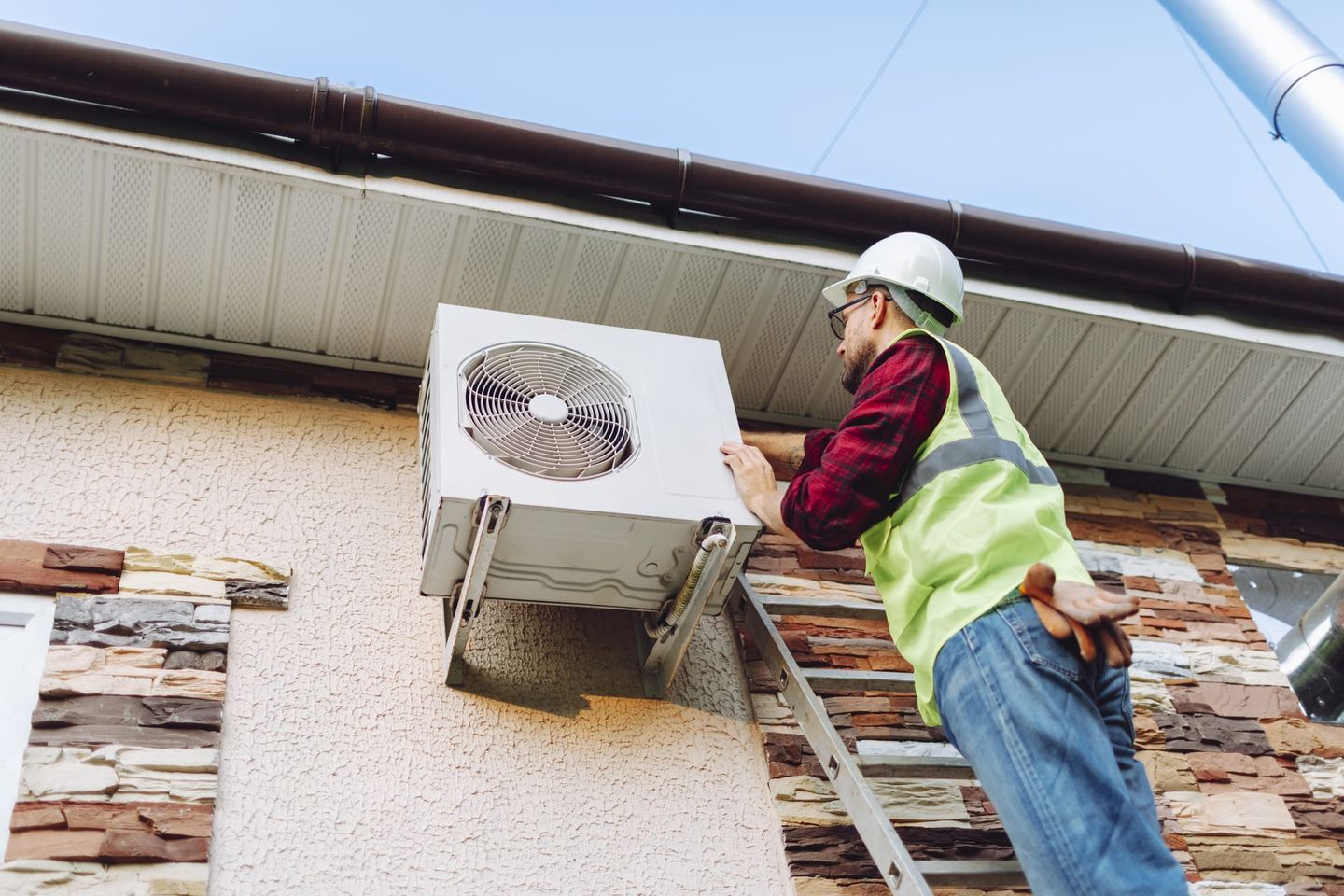 Handwerker mit Schutzhelm und Warnweste installiert eine Klimaanlage-Außeneinheit an der Fassade eines Hauses.