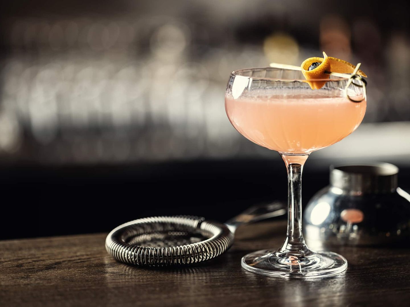 Ein rosa Cocktail im Glas mit Orangenzeste steht dekorativ auf einer Bar neben einem Barsieb. Getty