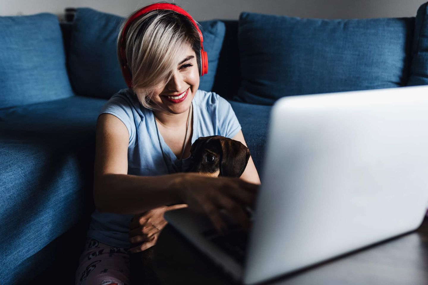 Junge Frau mit roten Kopfhörern sitzt zu Hause vor dem Laptop und arbeitet mit einem Hund auf dem Schoß Getty