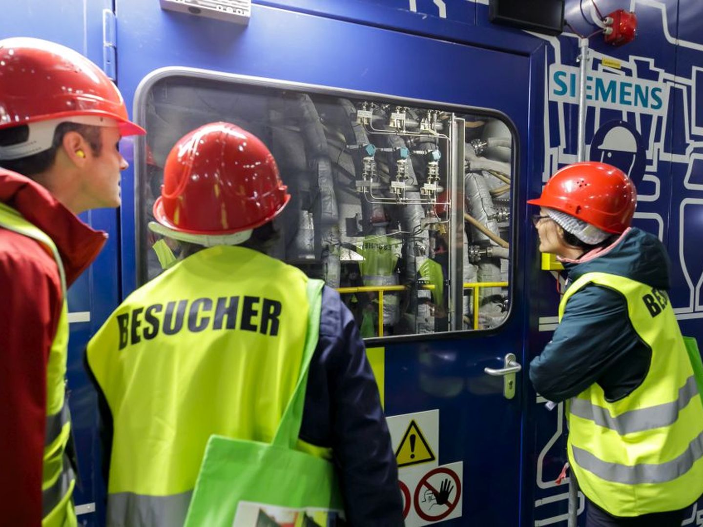 Besucher:innen mit Schutzhelmen und Westen betrachten Maschinen durch ein Fenster in einer Anlage. © Stadtwerke Düsseldorf © Stadtwerke Düsseldorf