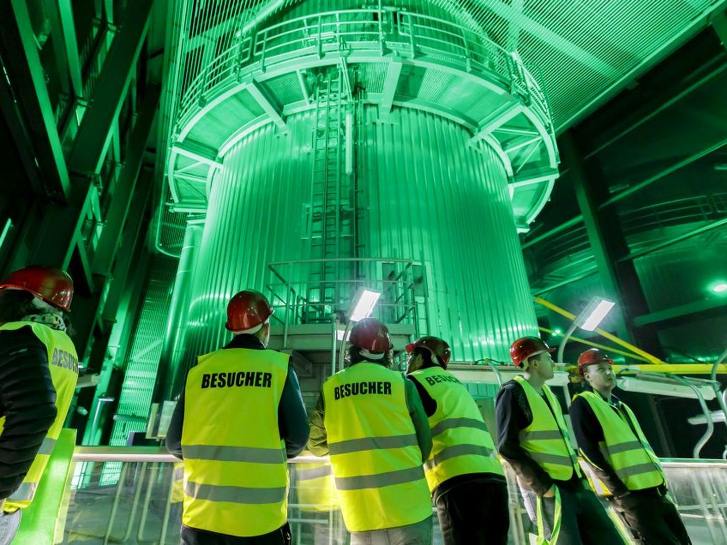 Besucher:innen mit Helmen und Westen betrachten ein großes, grün beleuchtetes Industriegebäude im Inneren. © Stadtwerke Düsseldorf © Stadtwerke Düsseldorf