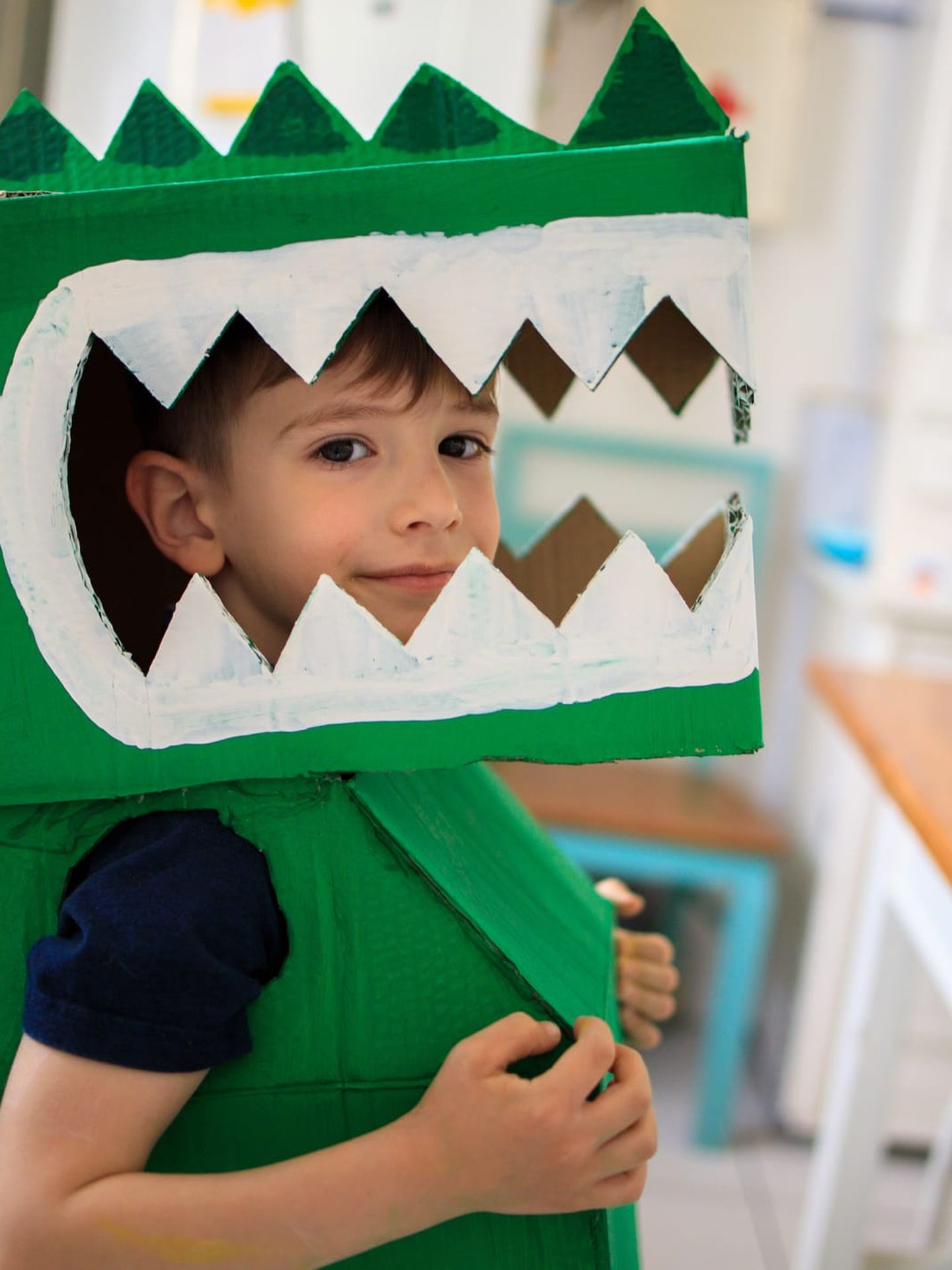 Ein Junge im selbst gebastelten grünen Dinosaurier-Kostüm aus Karton feiert fröhlich Karneval zu Hause