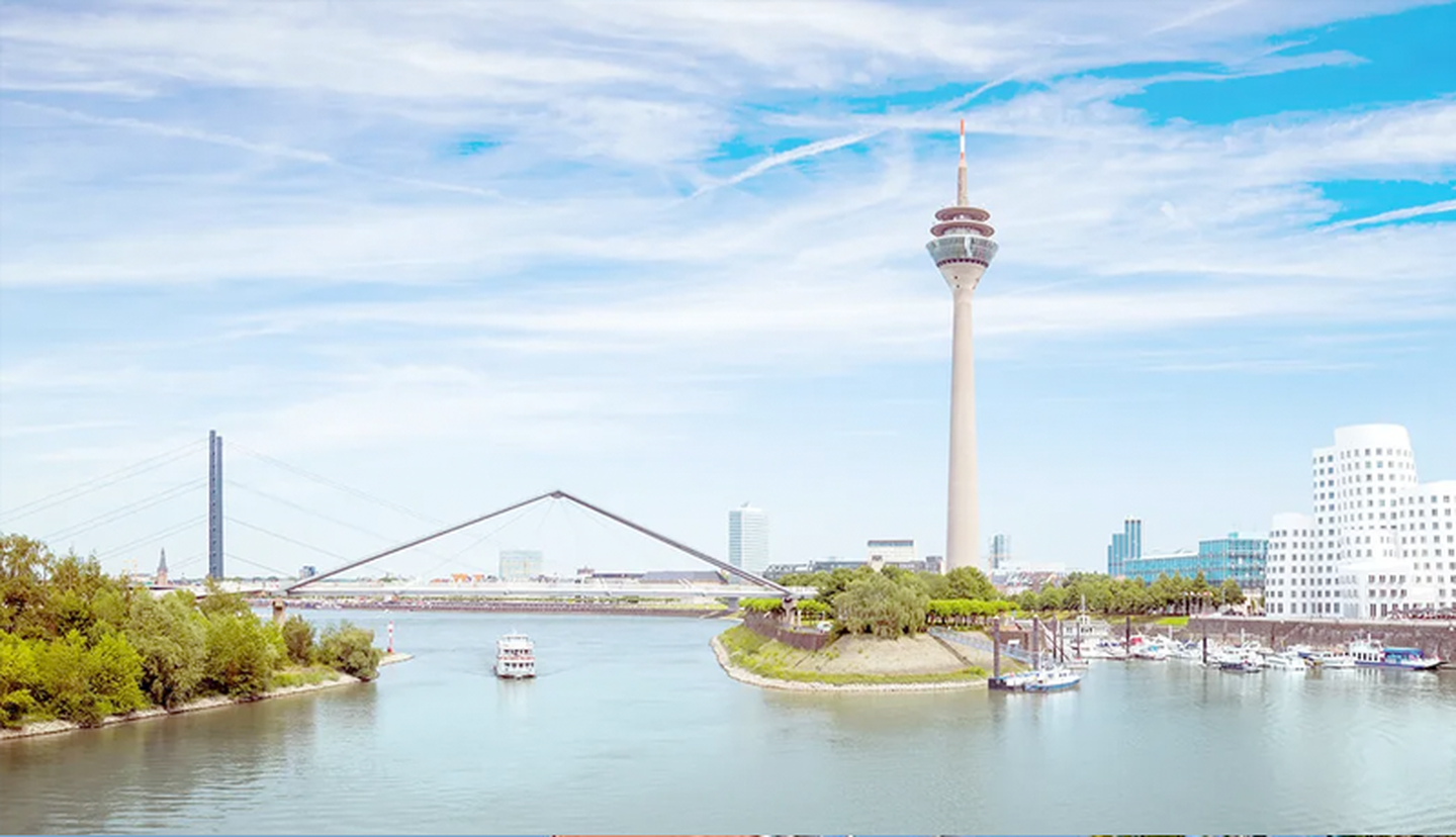 Flusslandschaft mit einem hohen Aussichtsturm, modernen Gebäuden, einer Brücke und Booten auf dem Wasser unter einem blauen Himmel mit vereinzelten Wolken.