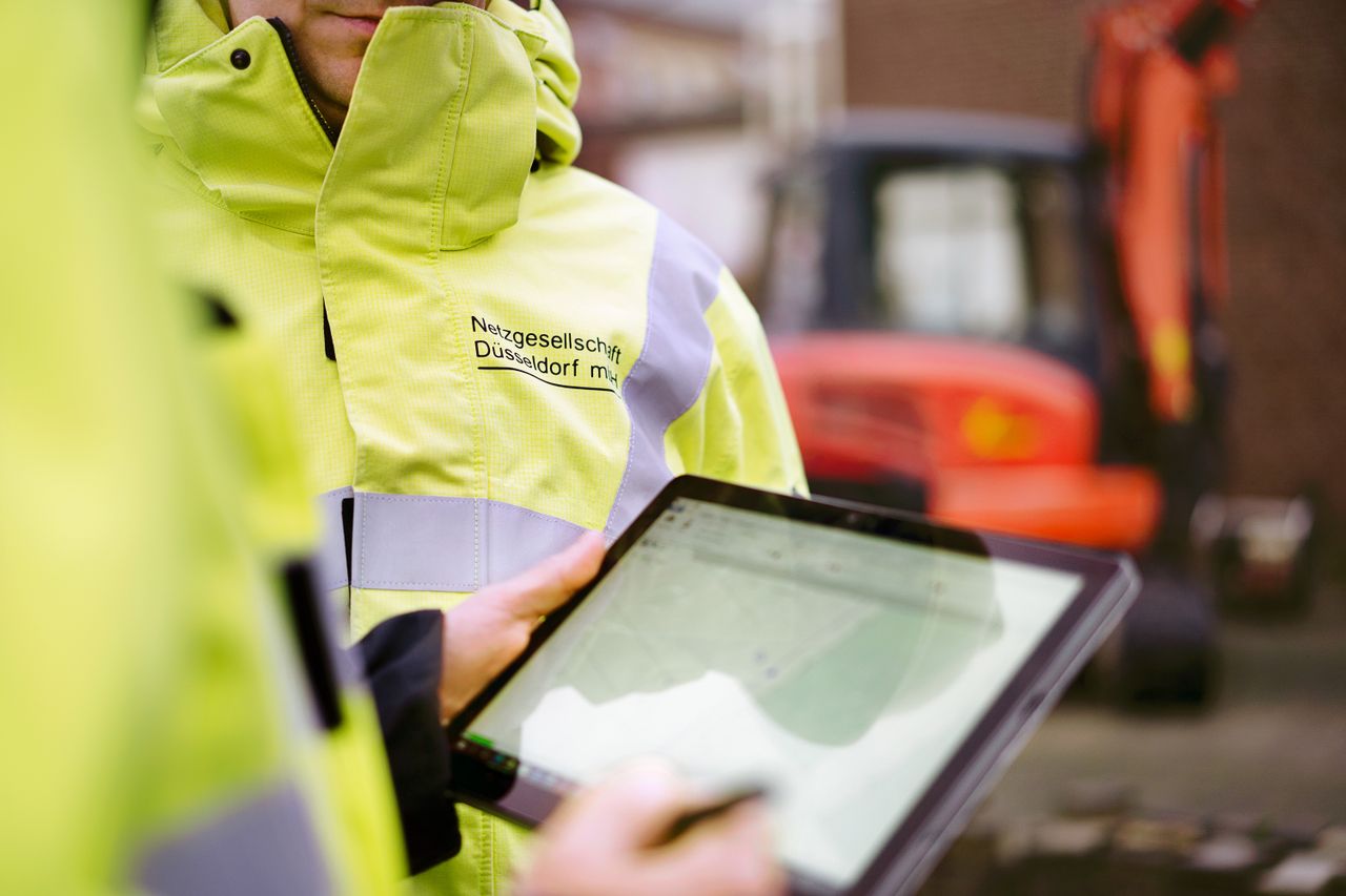 Worker in a yellow jacket uses a tablet outdoors, with an orange excavator blurred in the background.
