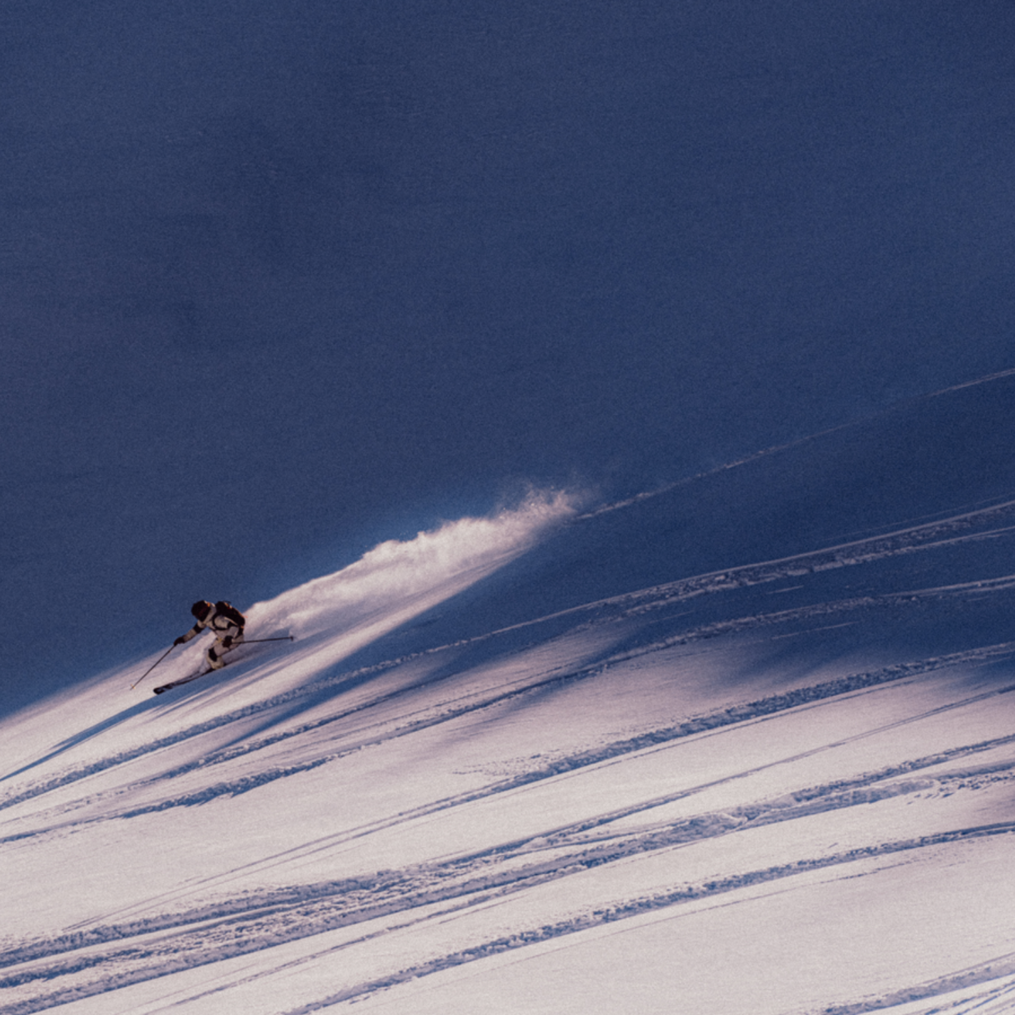 Skier carving through fresh powder on a sunlit, snowy slope against a clear blue sky.