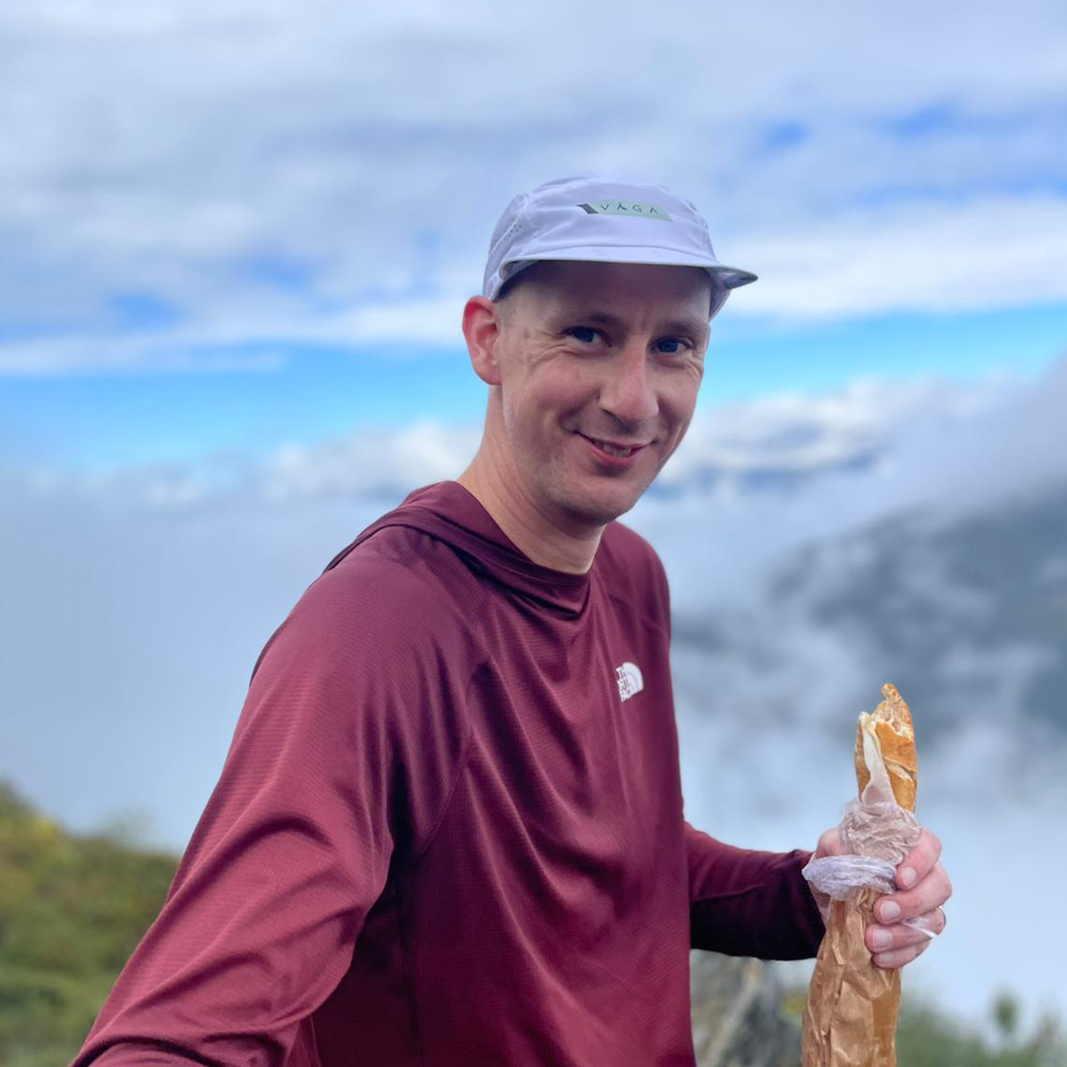 Man in a red long-sleeve shirt and white cap holding a baguette, standing on a foggy mountain with a cloudy sky backdrop.