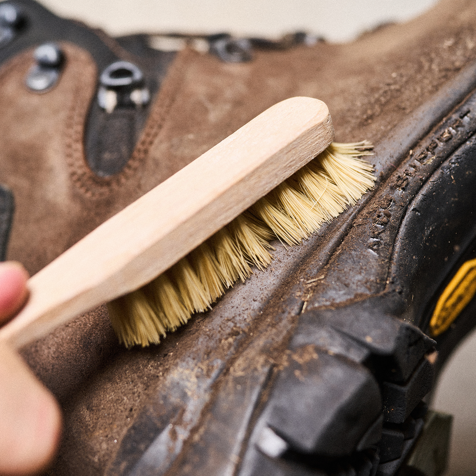 Close-up of a hand using a brush to clean a rugged brown hiking boot with black details and a textured sole.