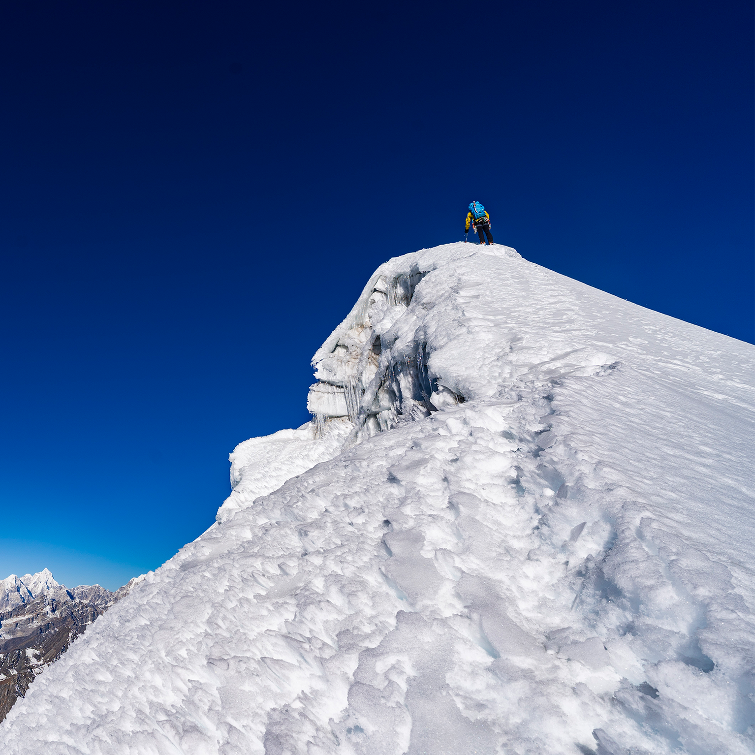A climber ascends a snowy mountain peak under a clear blue sky, with distant jagged peaks visible in the background.