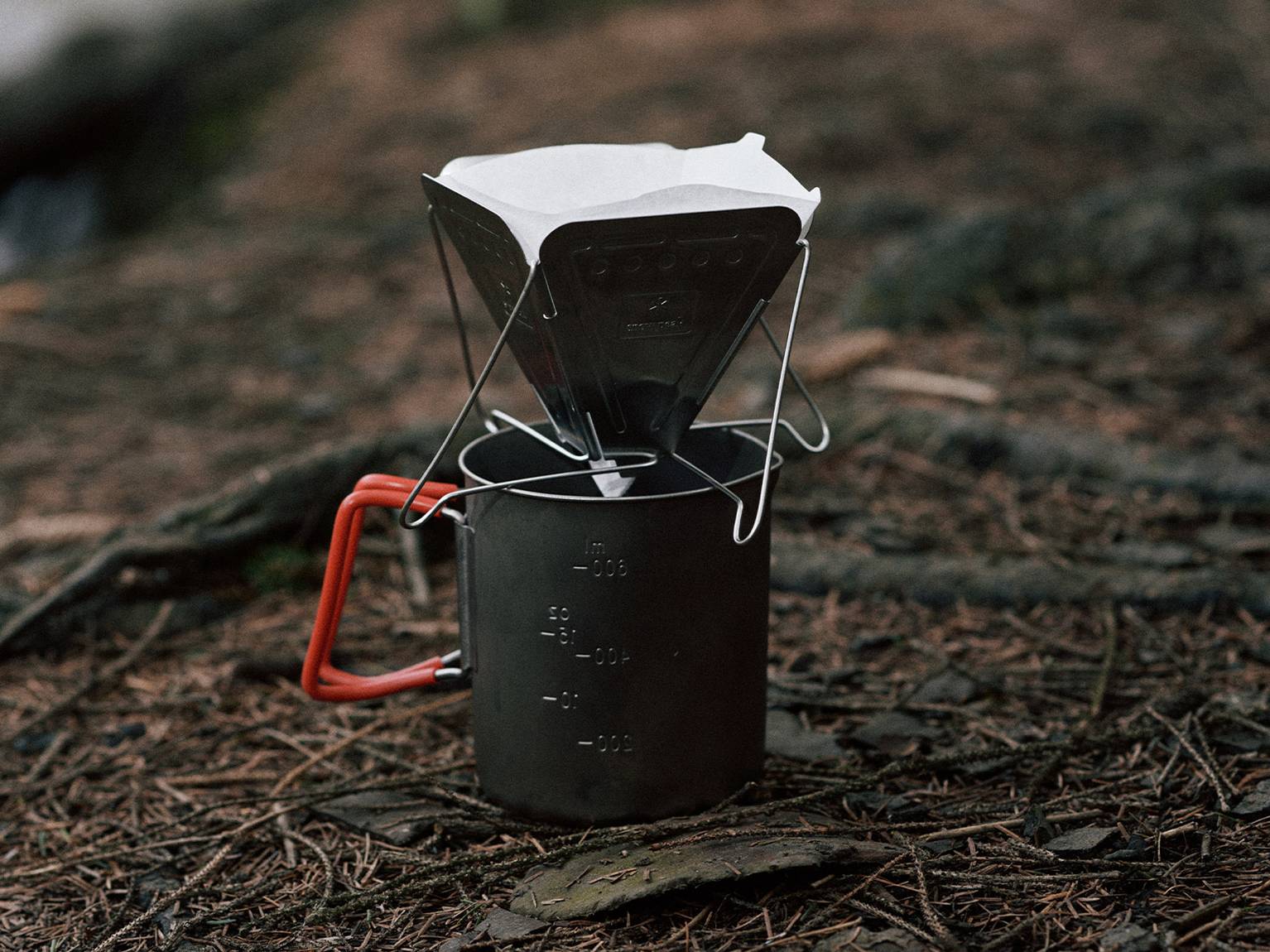 A portable coffee dripper on a metal cup resting on a forest floor, with a paper filter inside, surrounded by twigs and pine needles.