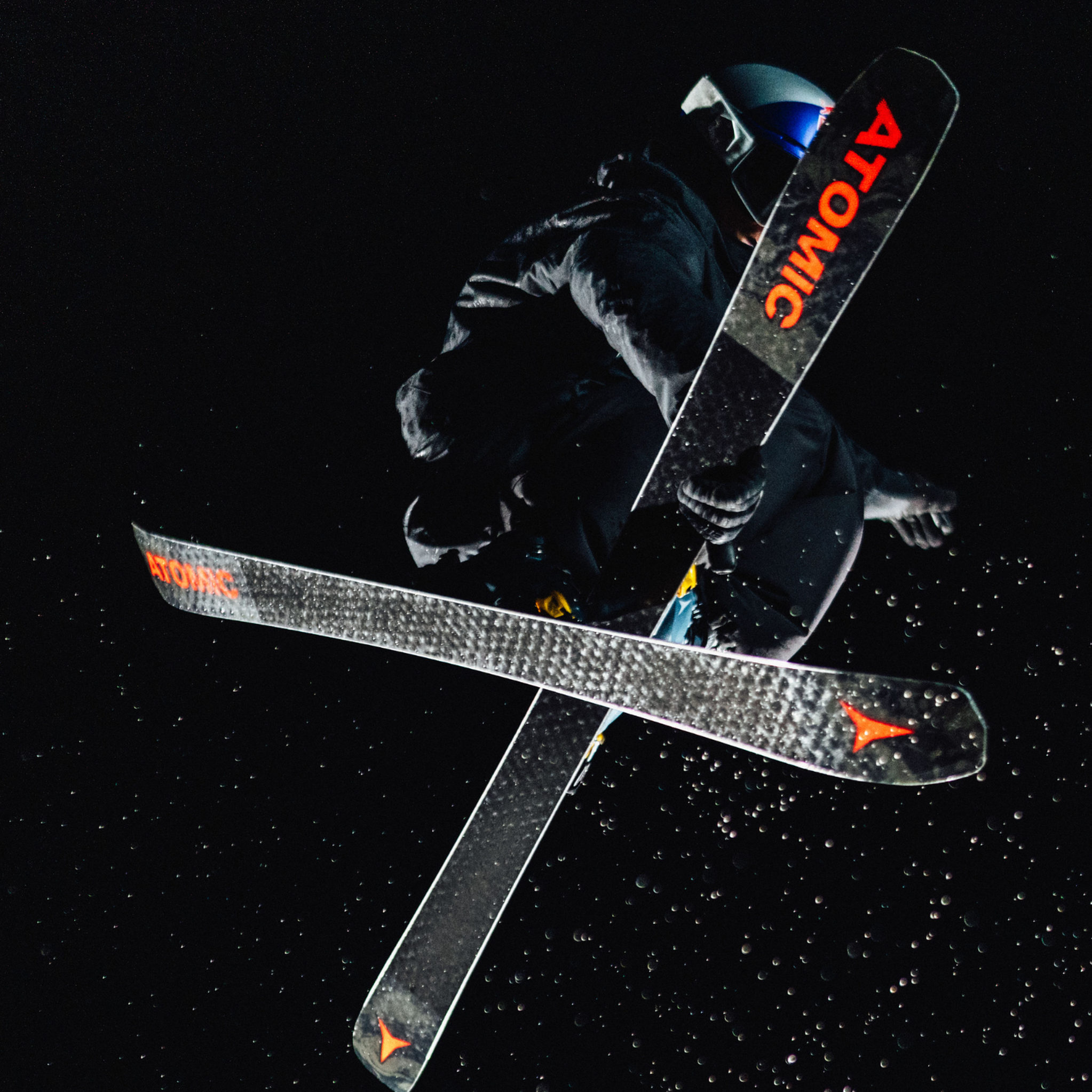 Skier performs a mid-air trick under bright lights at night, crossing skis with "Atomic" branding visible.