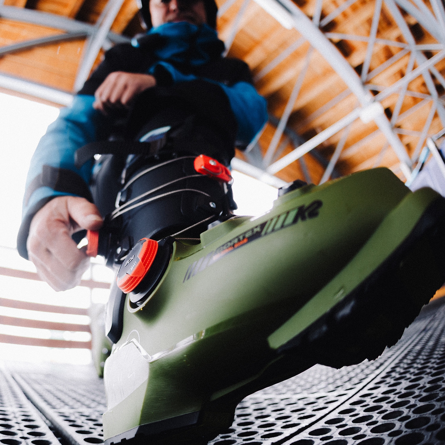 Close-up of a person adjusting a green ski boot with red buckles on a metal platform, under a wooden roof structure.