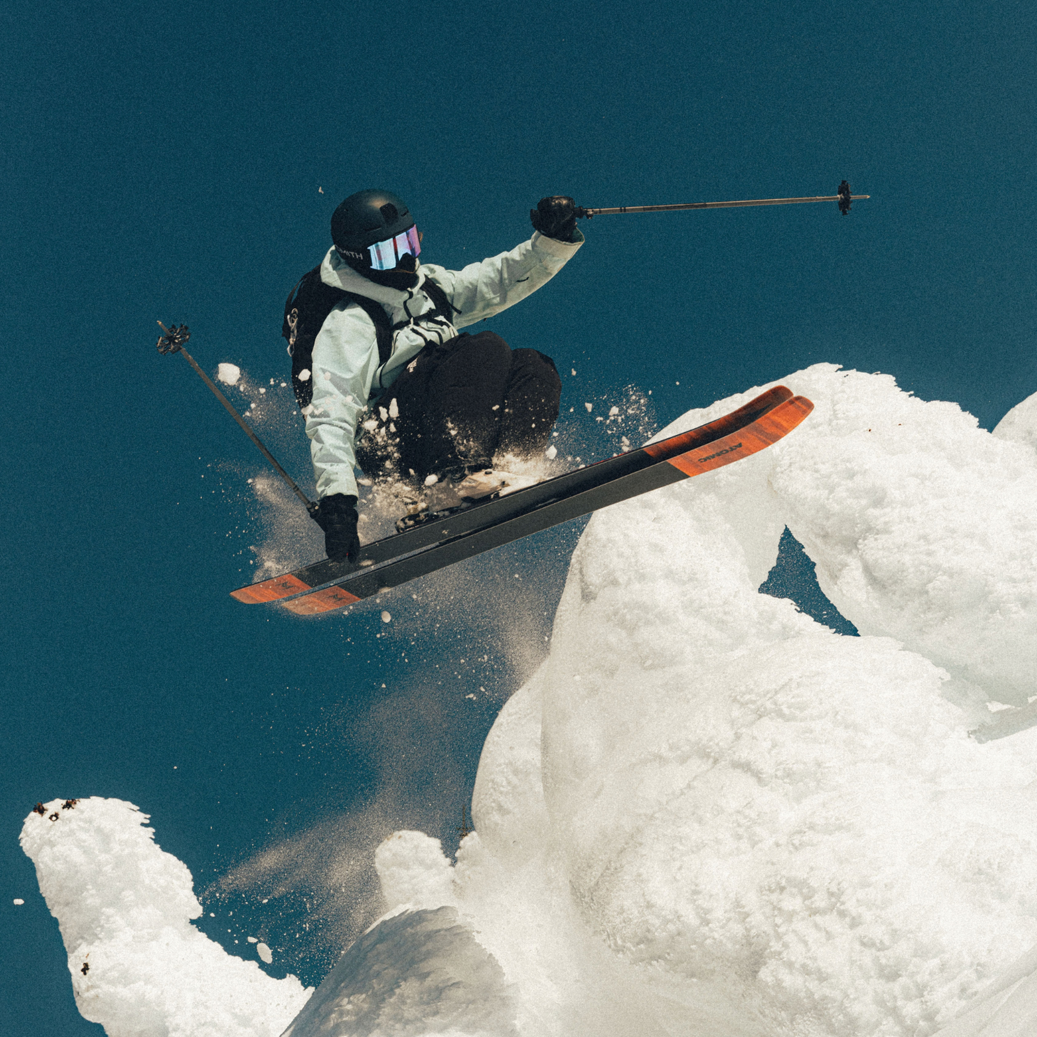 A skier in mid-air performs a jump off a snow-covered peak against a clear blue sky.