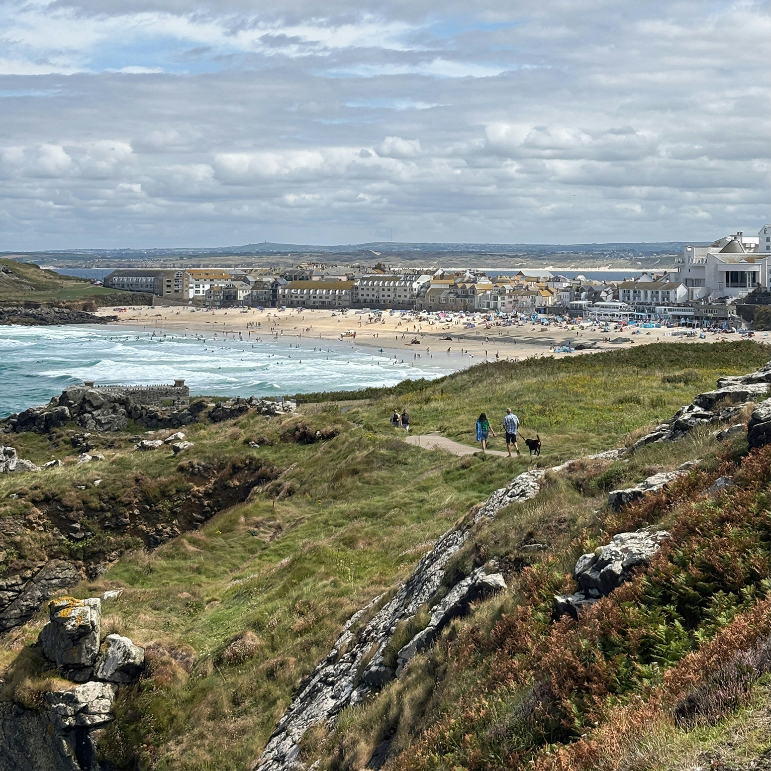 A coastal landscape with people walking along a grassy path. In the background, a beach and seaside town under a partly cloudy sky.