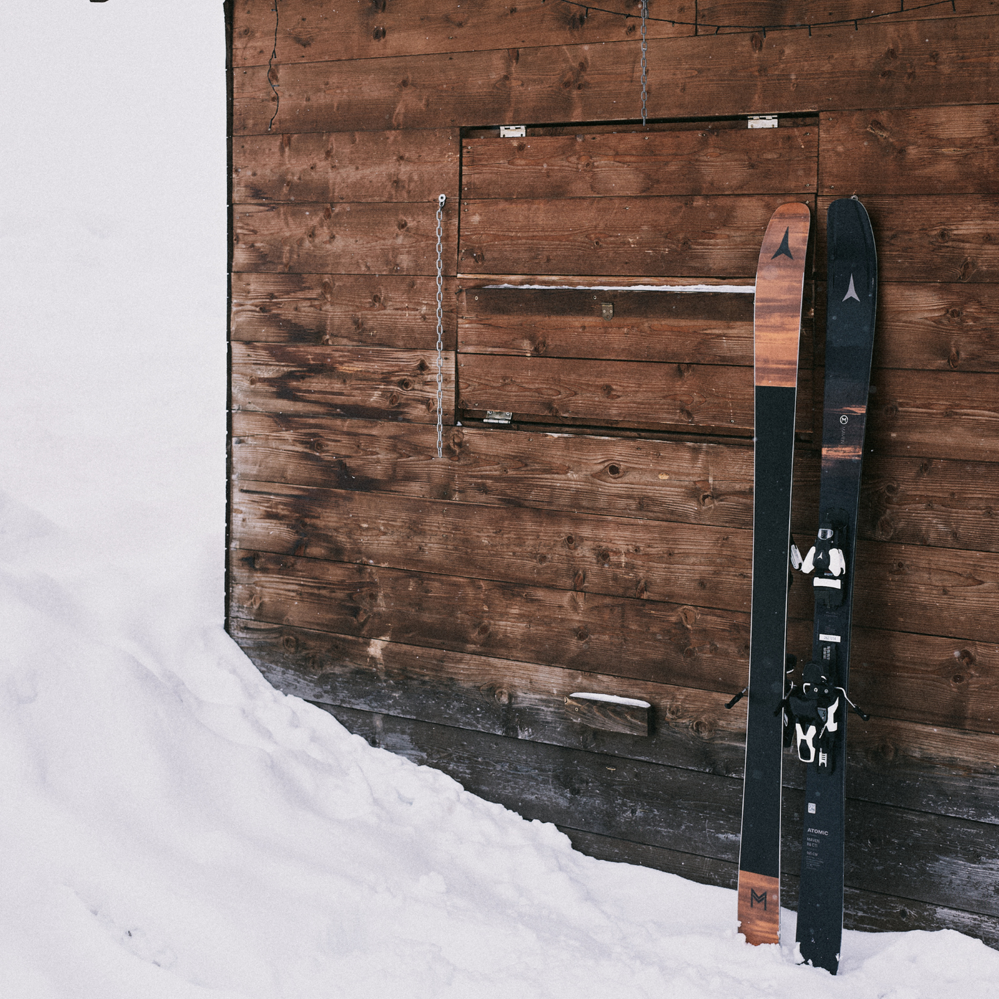 Two pairs of skis leaning against a rustic wooden wall, with snow covering the ground.