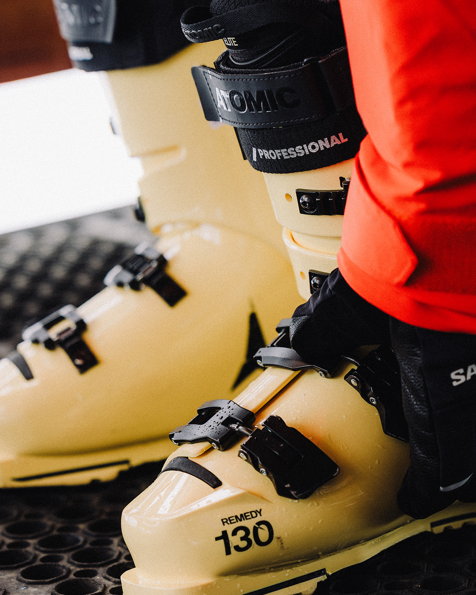Close-up of a person wearing yellow ski boots with black buckles, labeled "Atomic" and "Remedy 130," standing on a textured surface.