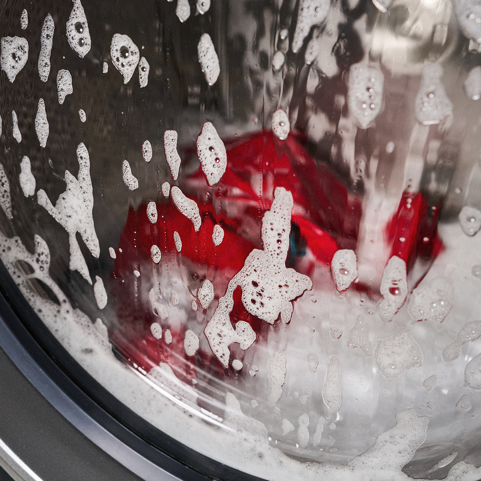 Close-up of a washing machine door with soapy water and bubbles on the glass, revealing red clothing being washed inside.