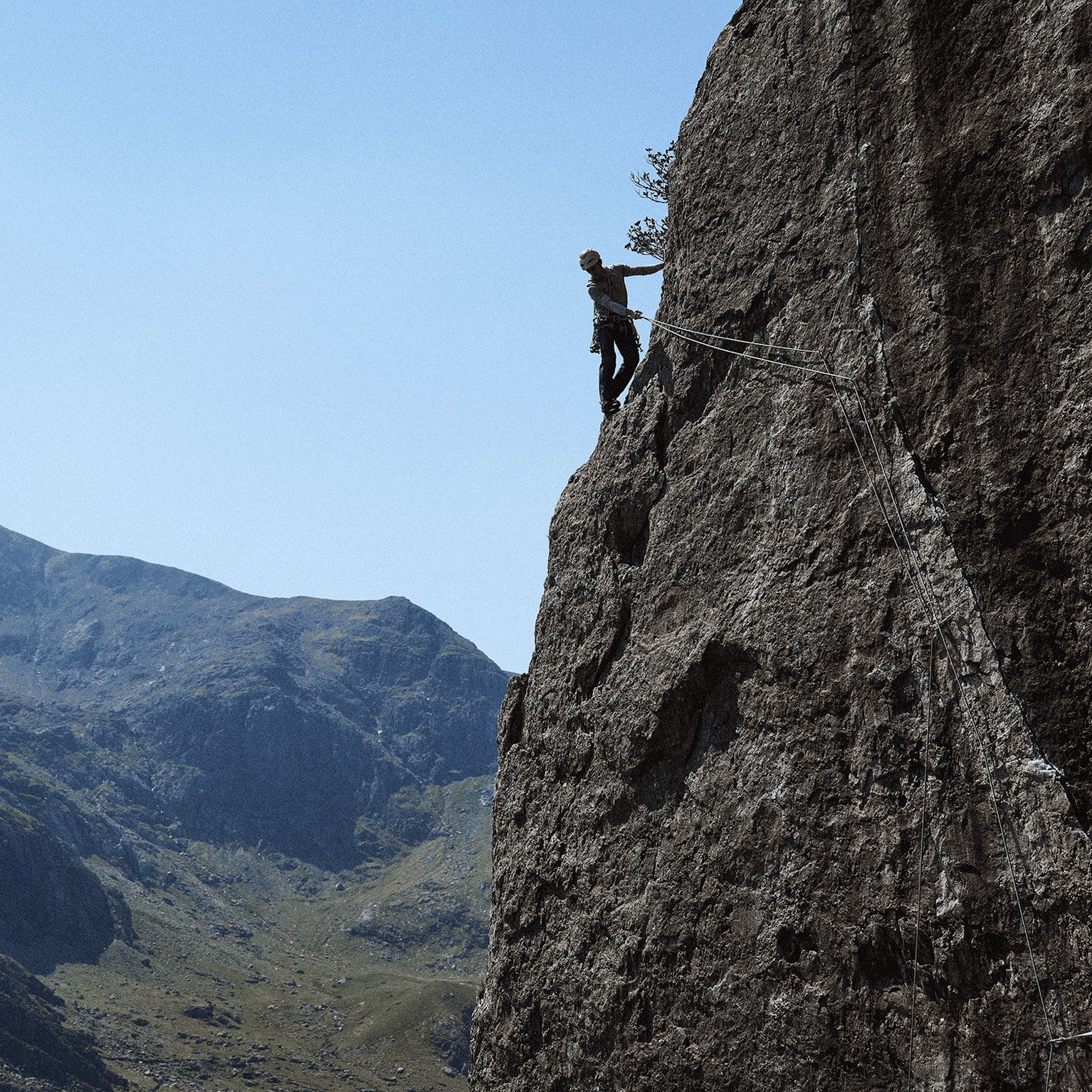 Rock climber scaling a steep cliff against a backdrop of rugged mountains and clear blue sky.