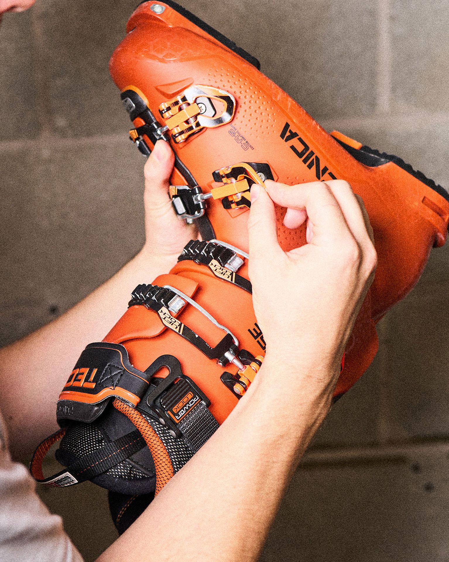 Person adjusting the buckles on an orange ski boot against a gray background.