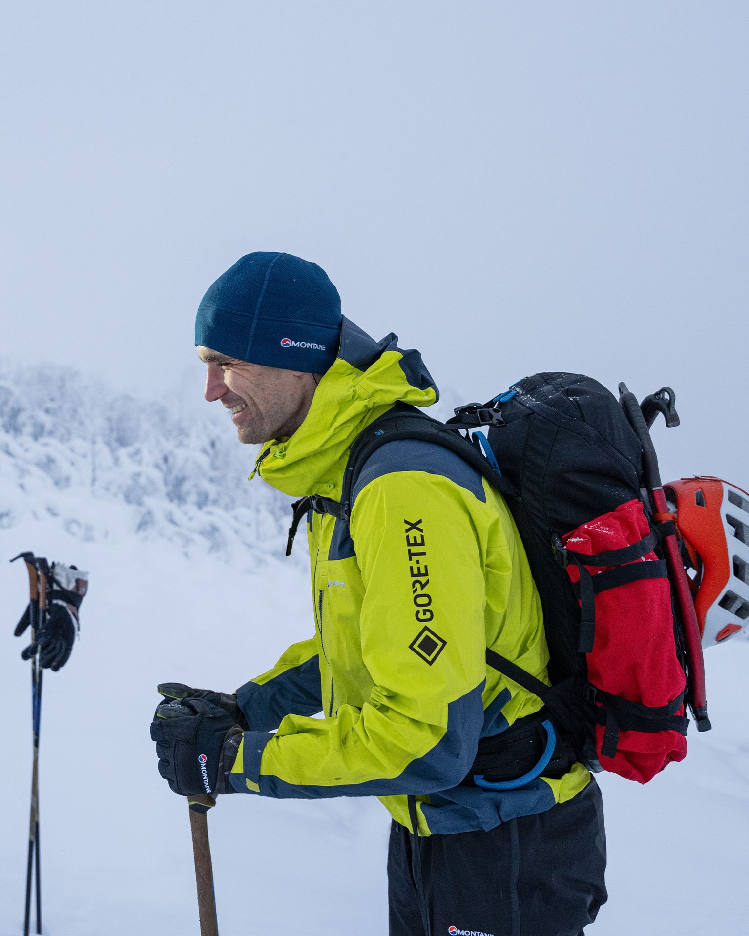 Smiling mountaineer in bright yellow GORE‑TEX jacket and blue beanie, carrying a rucksack with helmet and ice axe in snowy landscape