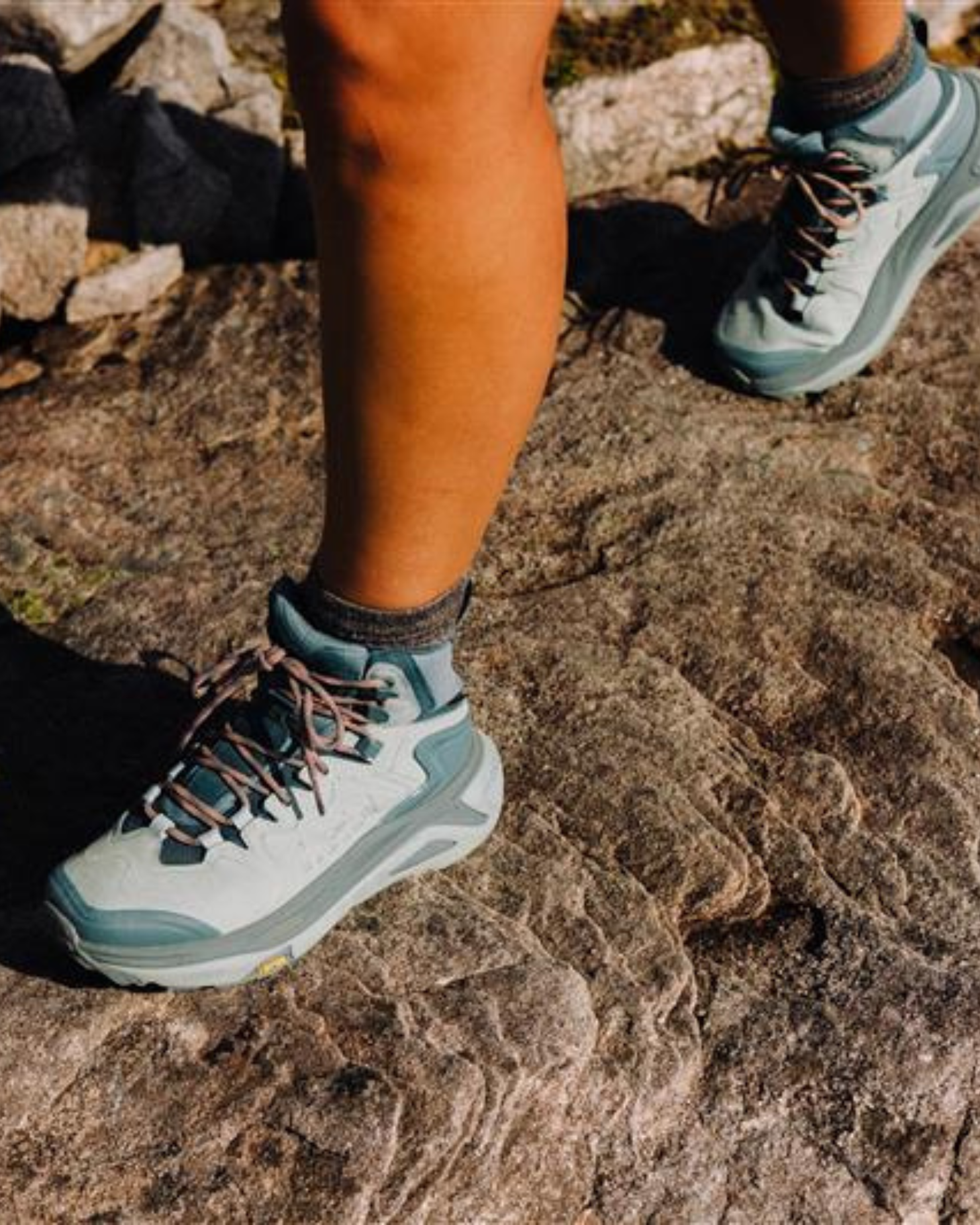 Close-up of a person wearing teal hiking boots standing on a rocky surface, showcasing durable footwear for mountain sports.