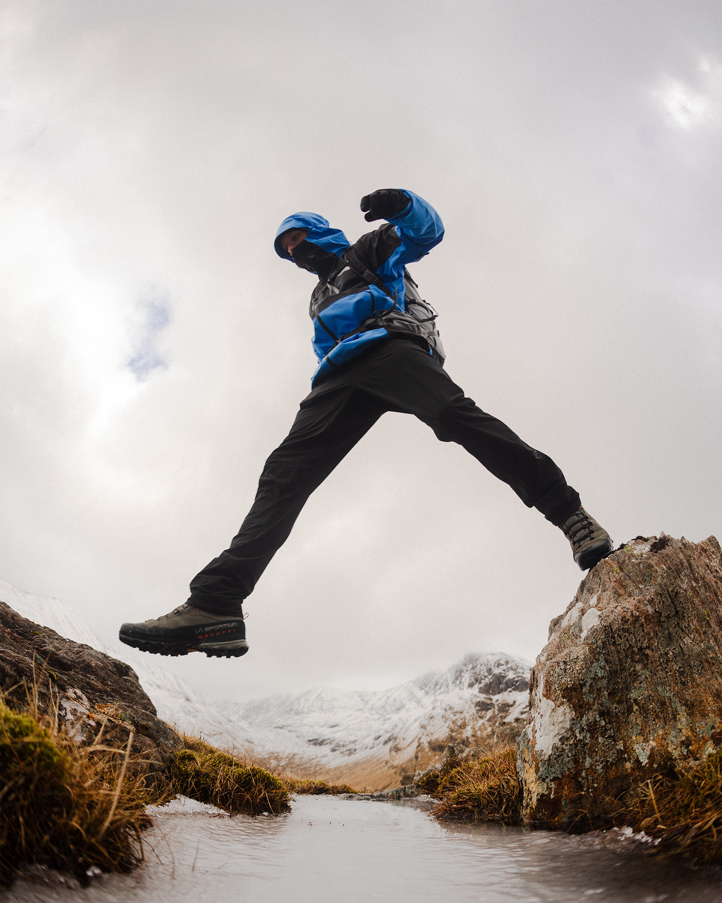 Person in blue jacket and black trousers leaps over a rocky stream in a mountainous landscape, wearing sturdy boots and a hood.