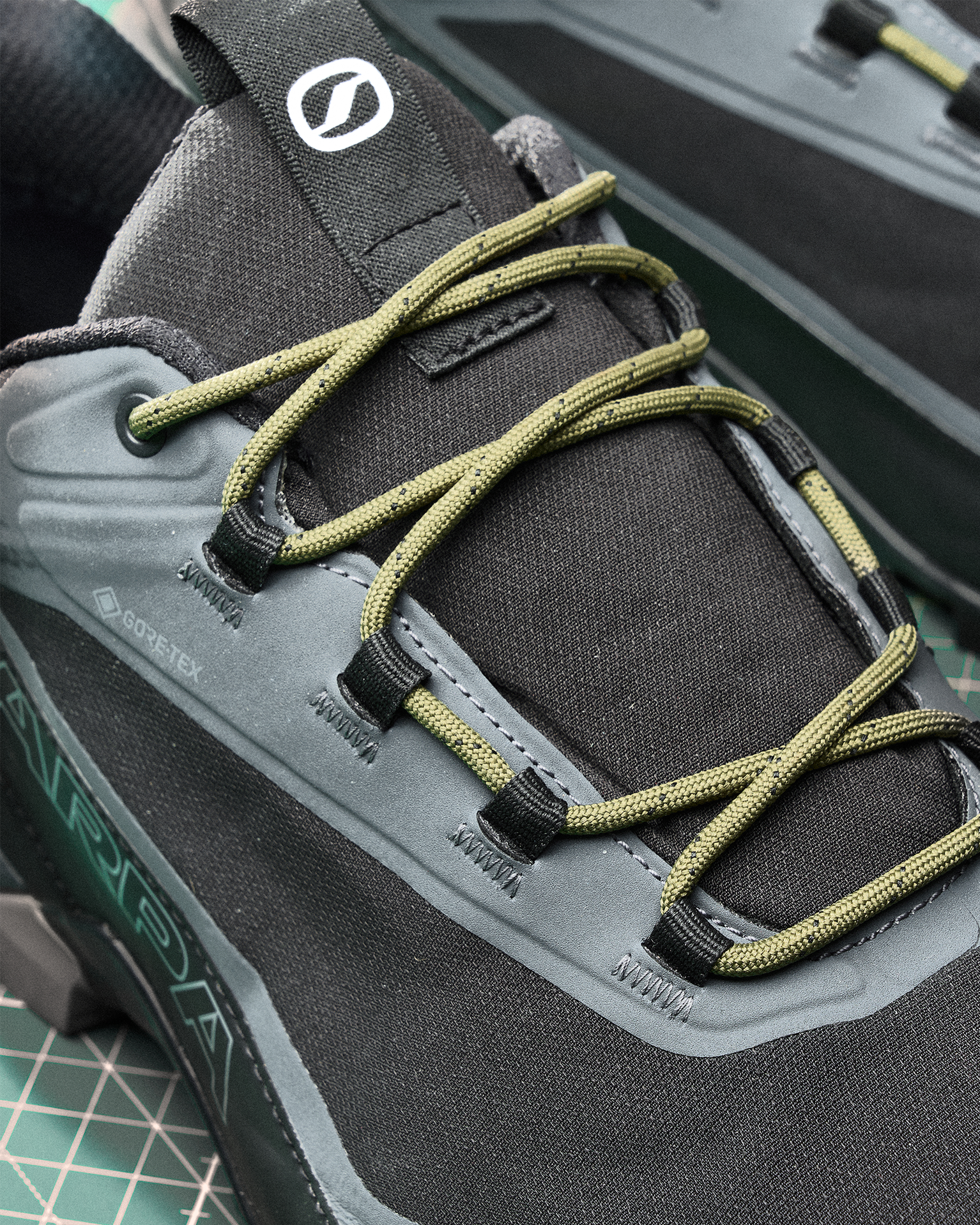 Close-up of black hiking shoes with green laces, featuring Gore-Tex material and a textured sole on a grid-patterned surface.
