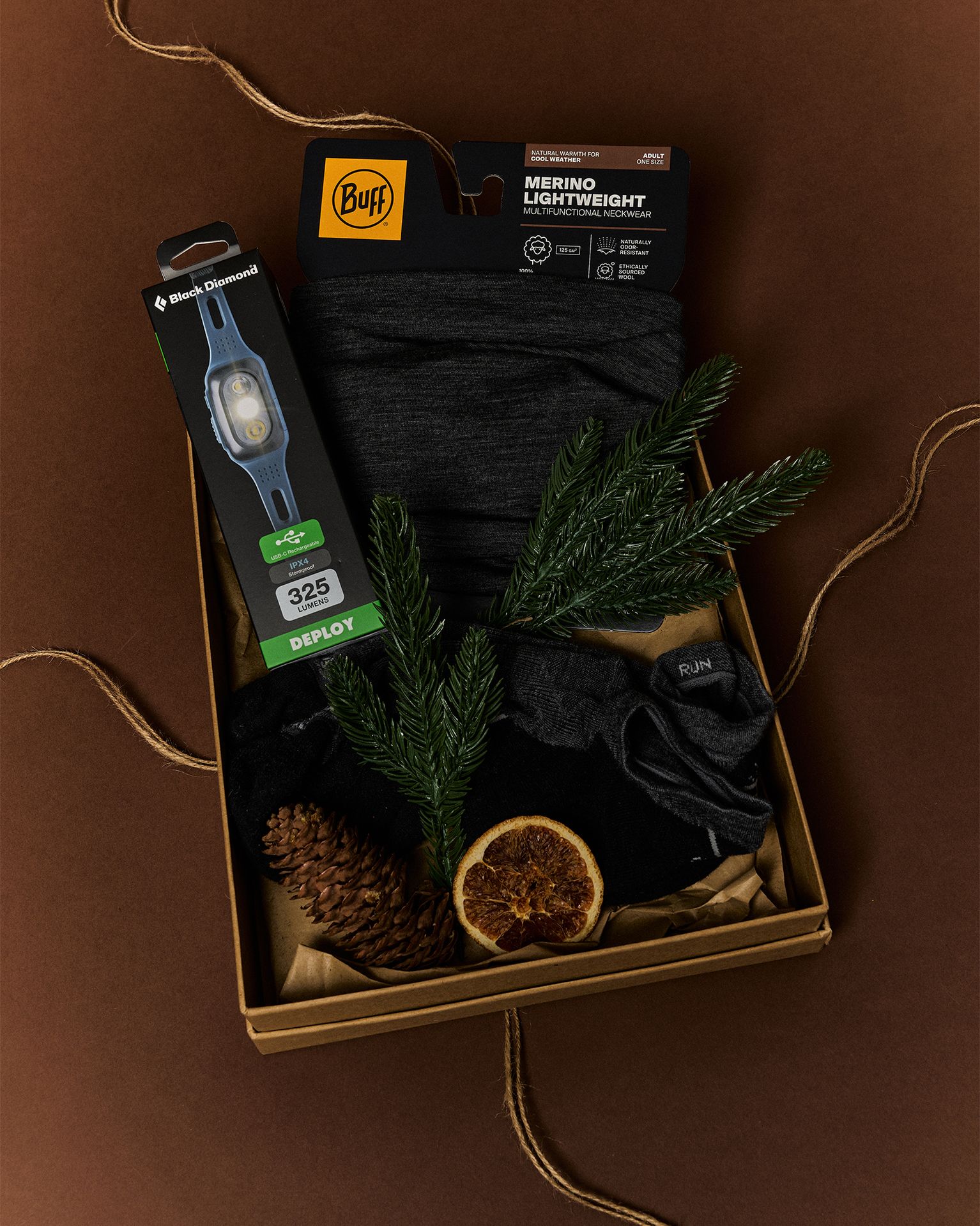 Gift box with merino wool clothing, a headlamp, pinecones, and a dried orange slice, arranged on a brown background.