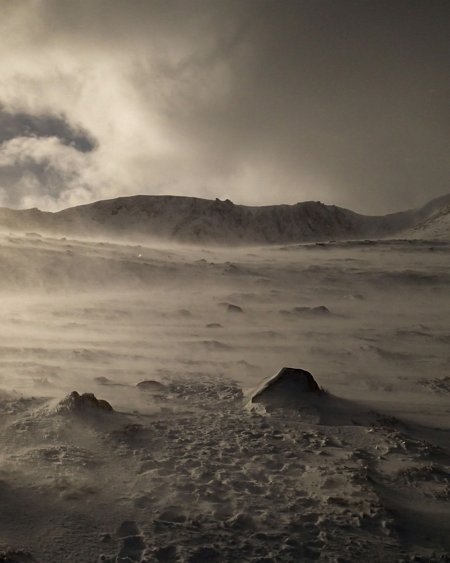 Snow-covered mountain landscape with strong winds creating snow drifts, under a cloudy sky. Rugged terrain with scattered rocks.