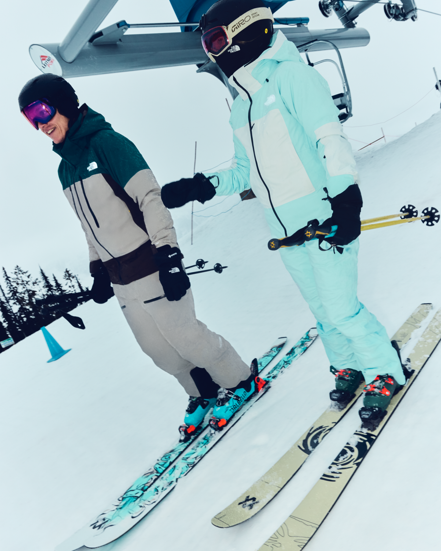 Two skiers in stylish gear smiling as they glide down a snow-covered slope near a ski lift.