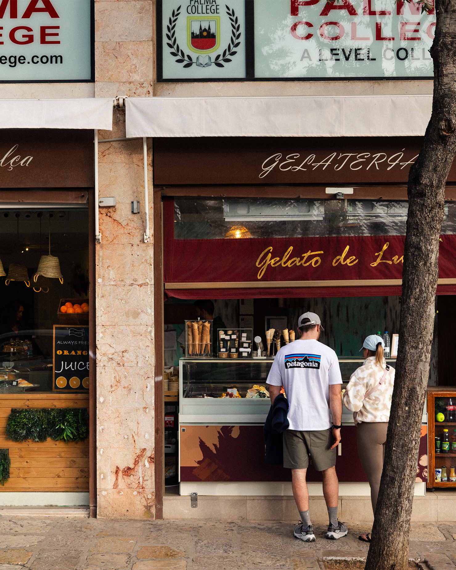 A couple stands in front of a gelato shop, Gelato de Luxe, deciding on flavours. The shopfront has a rustic design with a tree nearby.