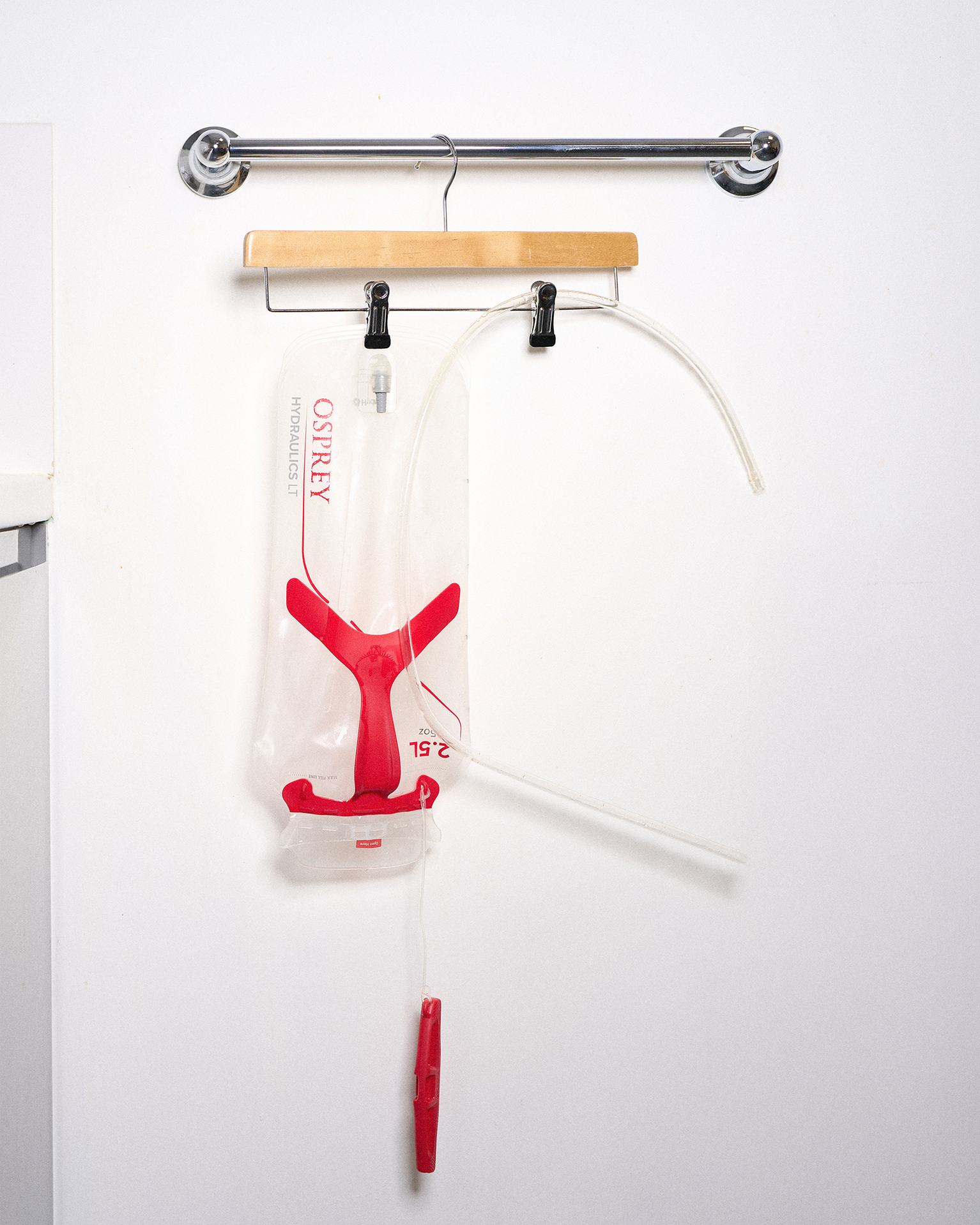 A hydration reservoir hangs from a wooden hanger on a metal rod, with tubes and clips attached, against a white wall.