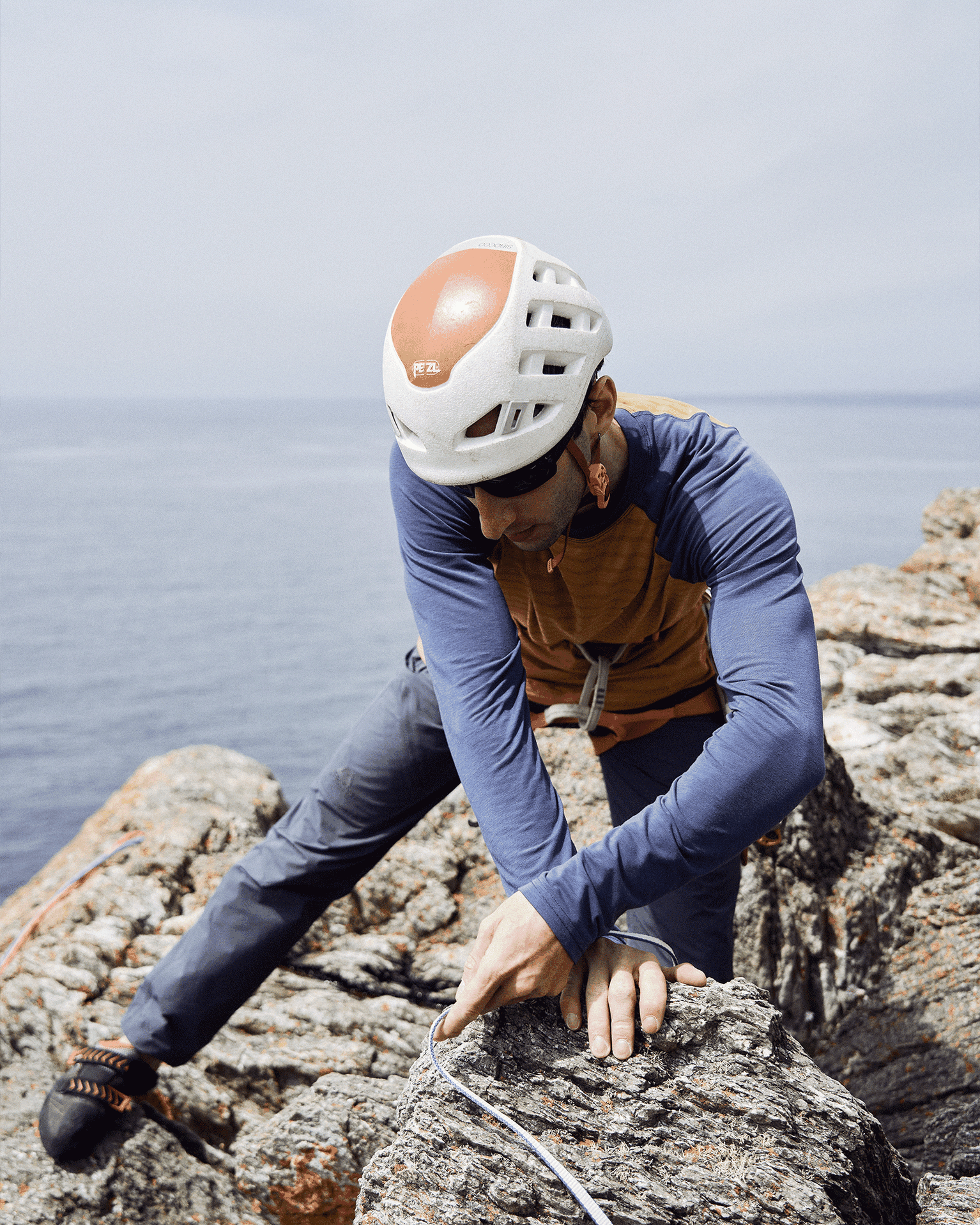 Climber in helmet gripping rope on rocky cliff by the sea, demonstrating focus and expertise.