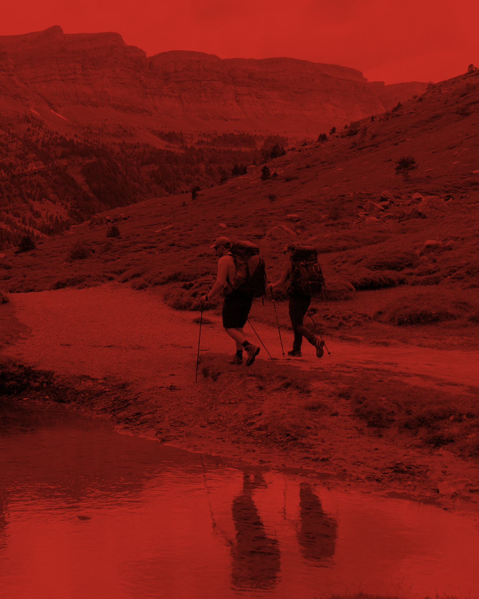 Two hikers with backpacks traverse a mountain path beside a reflective pool, with rugged peaks in the background under a red hue.