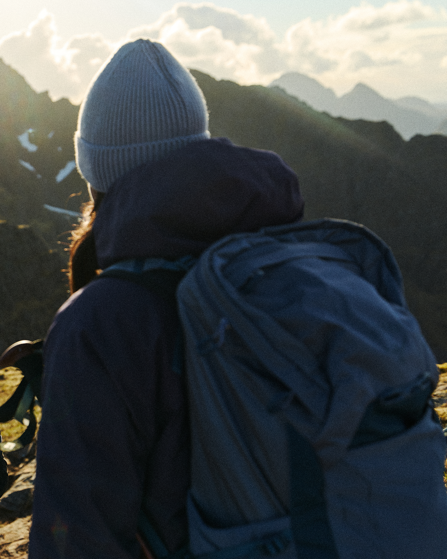 Person in a beanie and jacket with a large backpack, looking at a mountainous landscape during a sunrise or sunset.