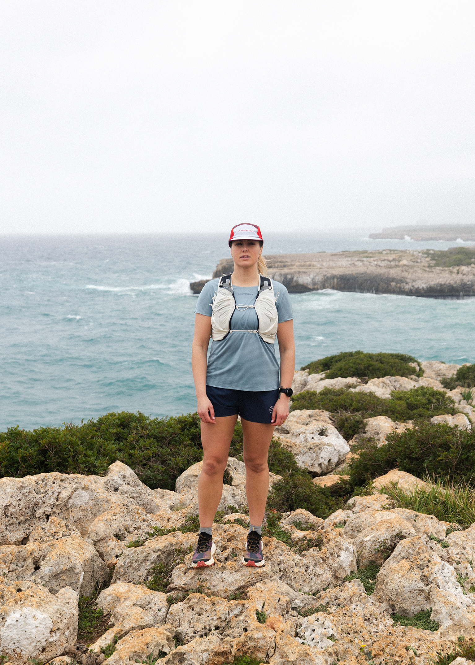 A person in sportswear stands on rocky terrain by the sea, wearing a cap and a running vest, with a cloudy sky in the background.