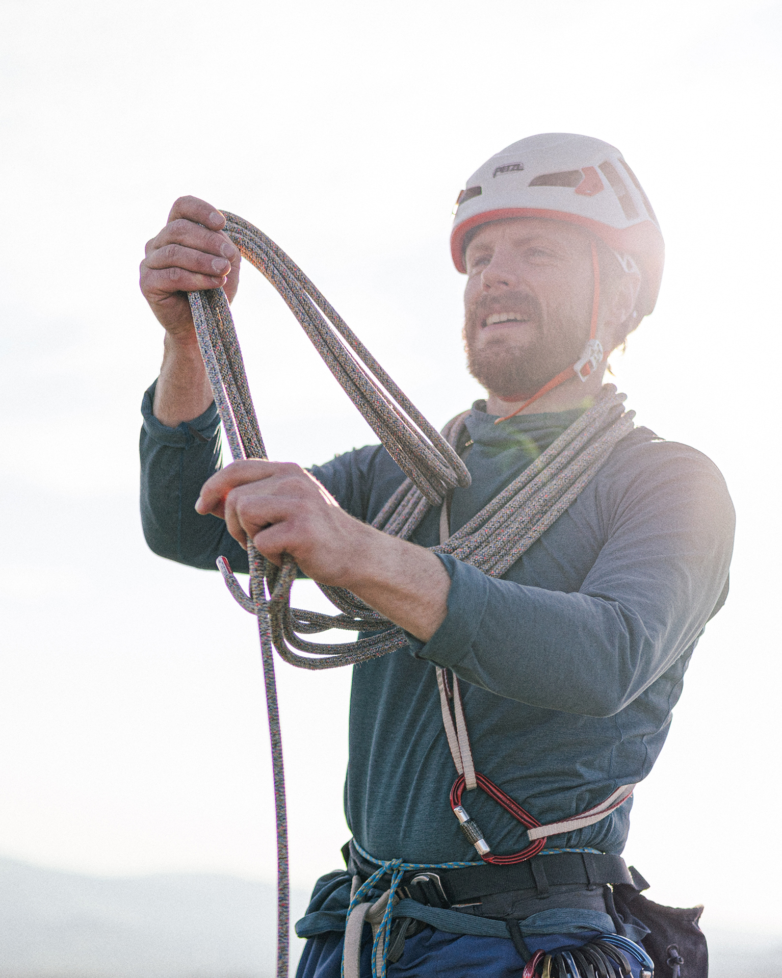 A climber wearing a helmet and harness manages a coiled rope outdoors, with a bright sky in the background.
