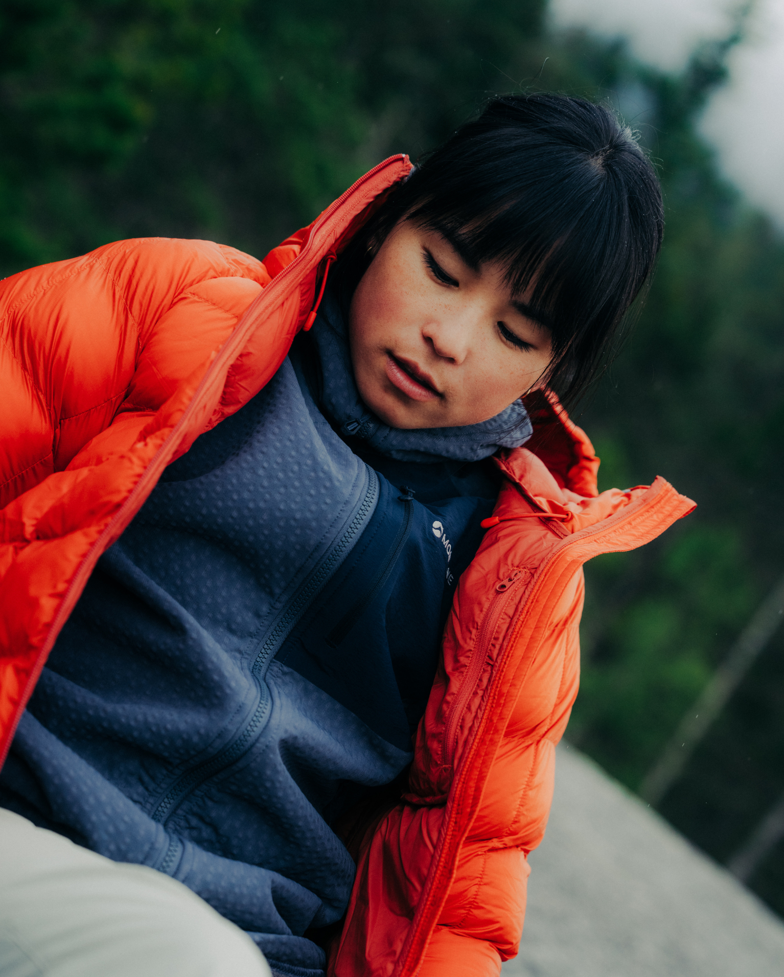 A woman in a Montane red Icarus down jacket over a blue fleece looks down thoughtfully, set against a blurred outdoor background.