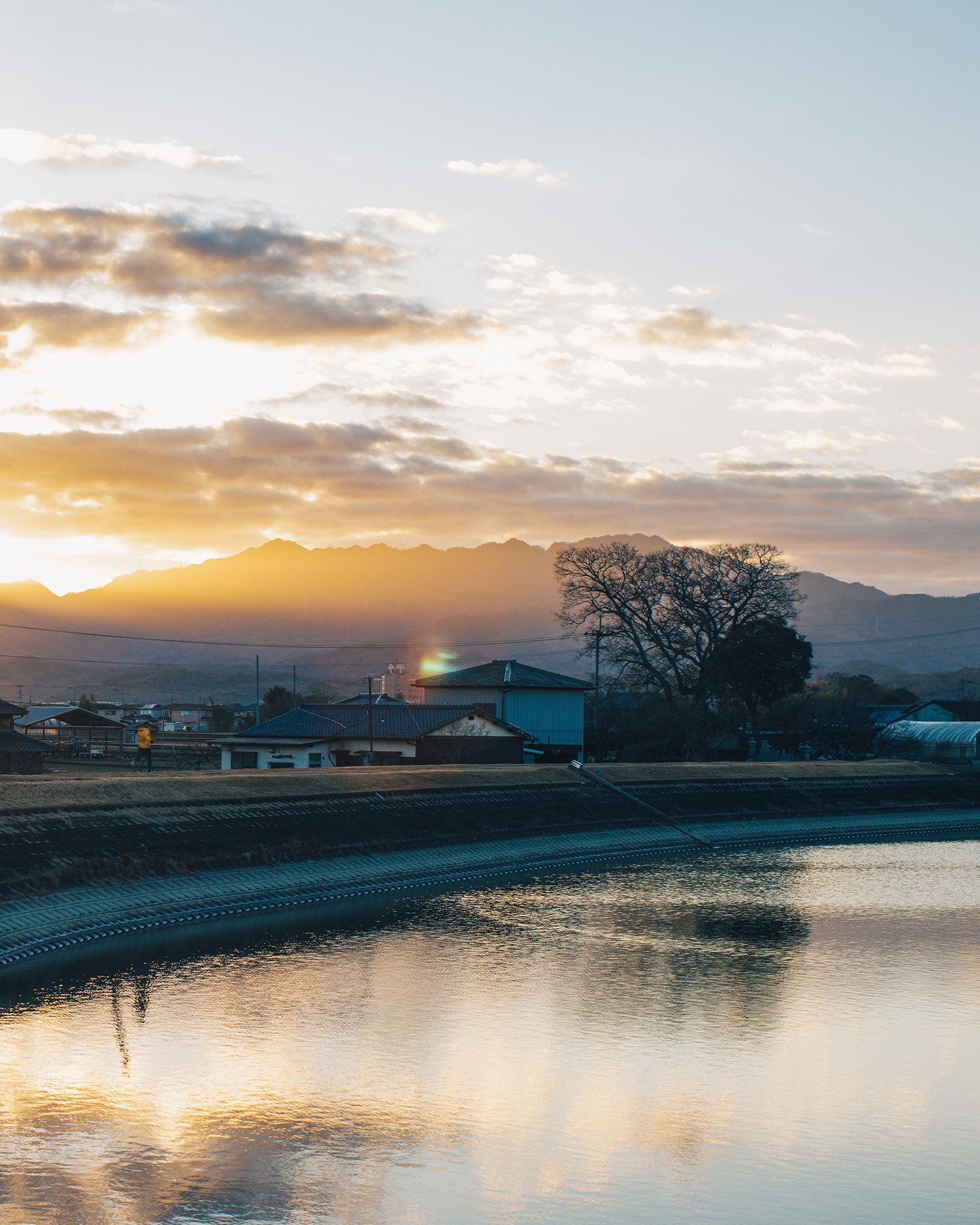 Sunrise over distant mountains, reflecting on a calm river with a tree and buildings silhouetted in the foreground.