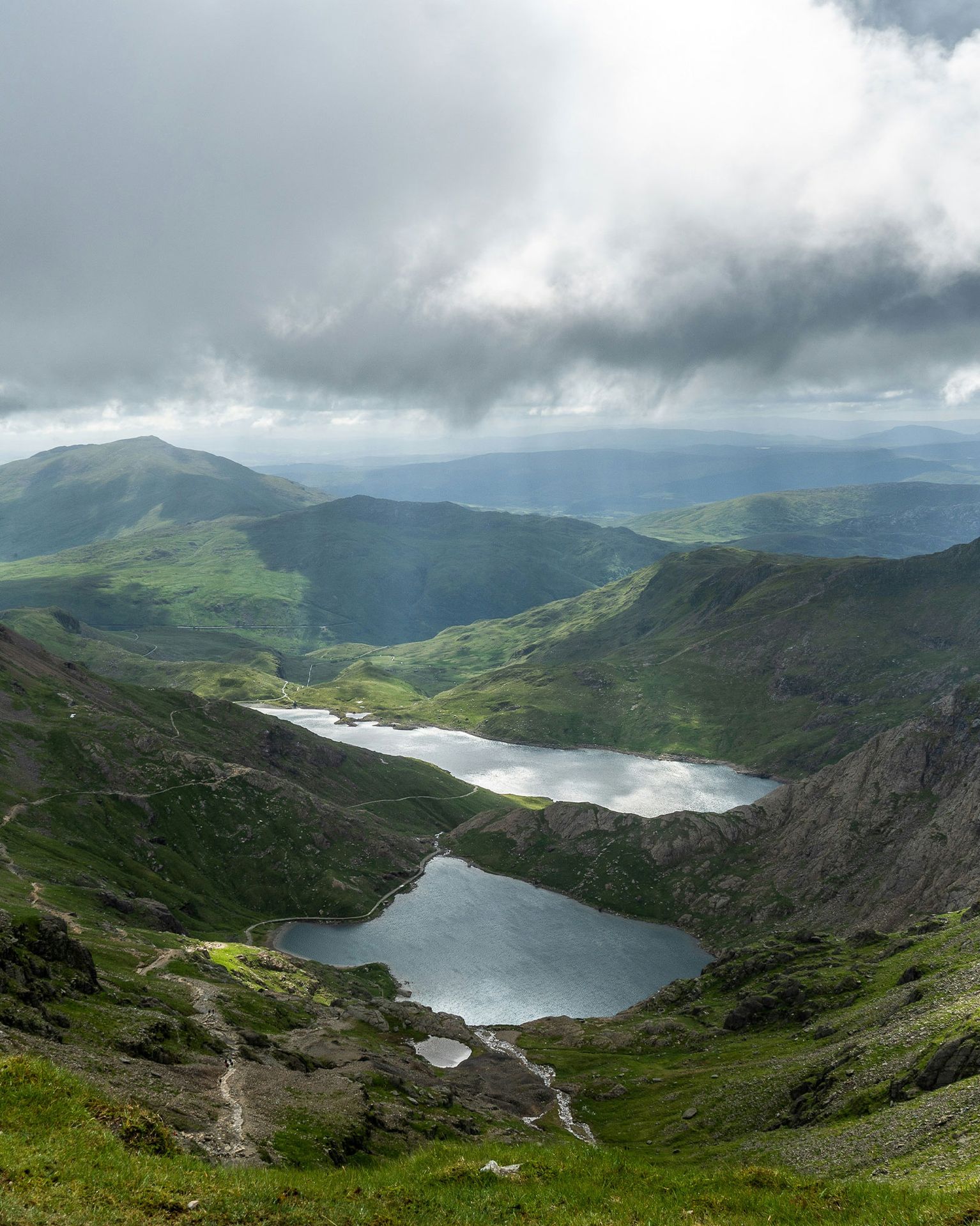 Mountain landscape with two serene lakes, green valleys, and dramatic clouds casting shadows over the scene.