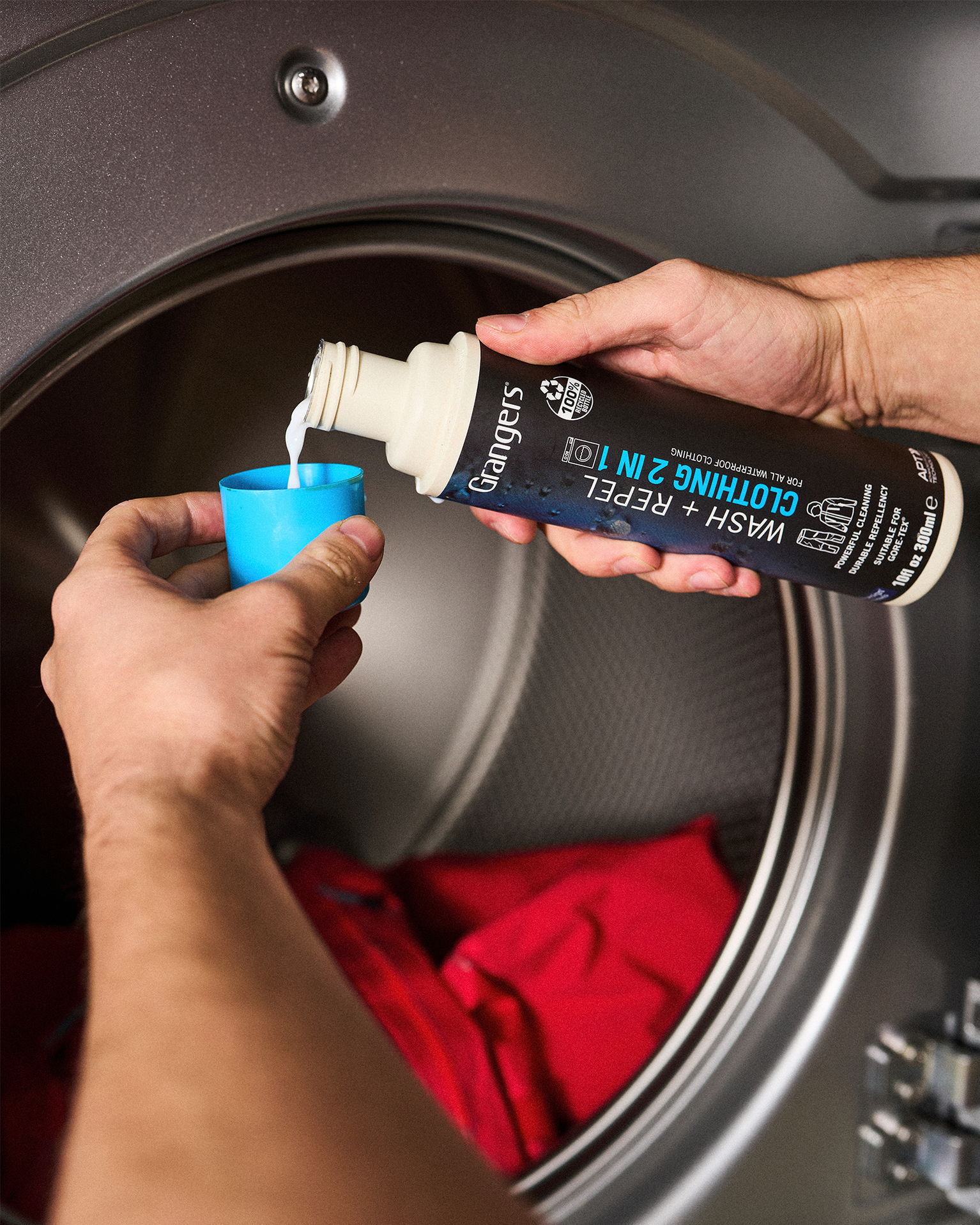 Person pouring Grangers clothing wash into a blue cap in front of an open washing machine with red clothes inside.