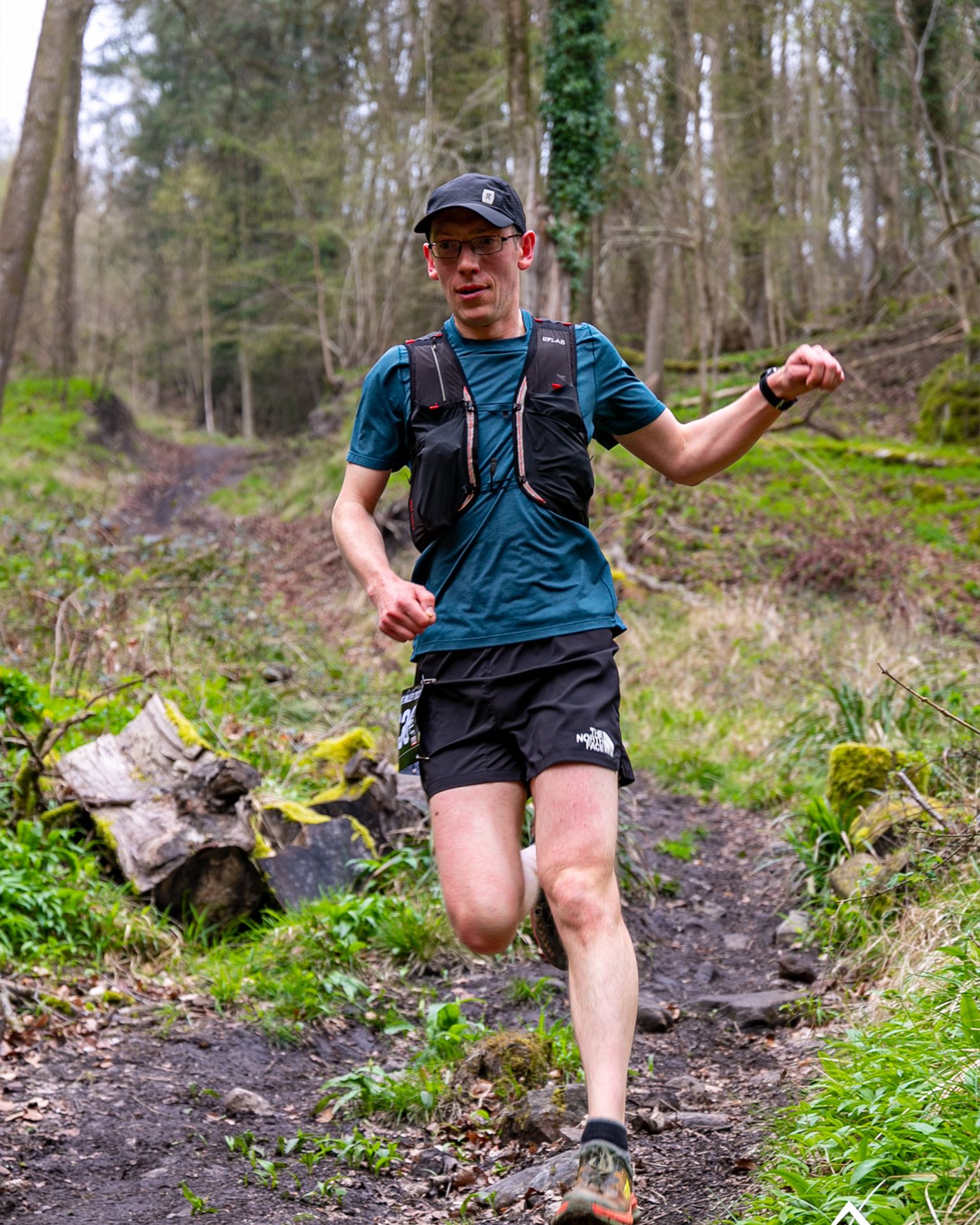 Runner in a forest wearing a hydration vest, blue shirt, and black shorts, navigating a rugged trail with vibrant greenery surrounding.