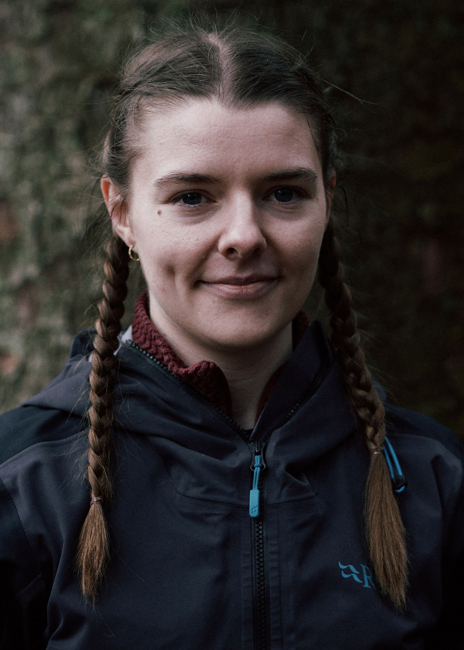 Woman with braided hair, wearing a dark jacket, stands outdoors with trees in the background, looking directly into the camera.