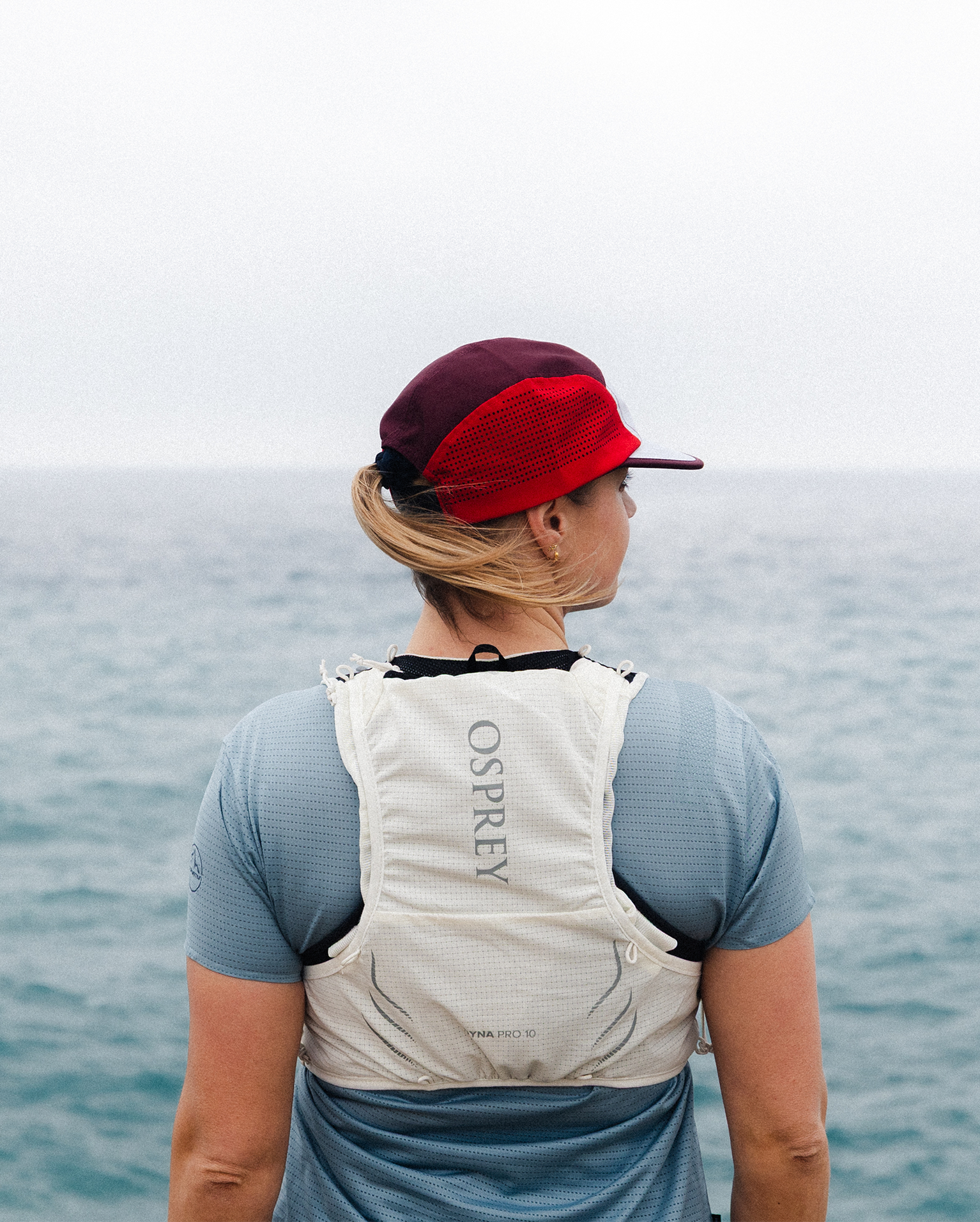 Person wearing a white Osprey hydration pack stands by the sea, wearing a red cap and blue shirt, gazing out at the water under a cloudy sky.