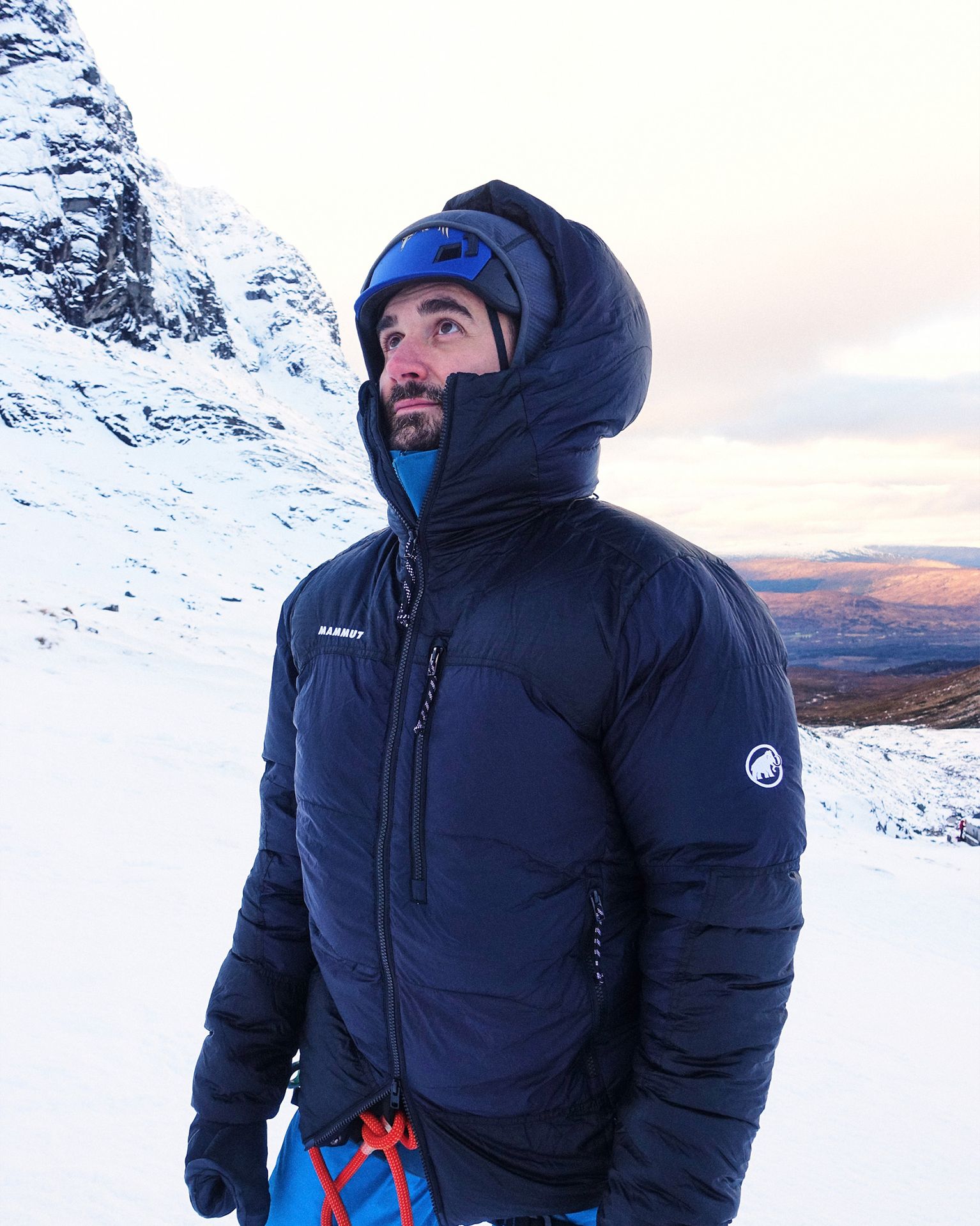 Man in a warm, dark jacket and blue helmet standing in snowy mountains, looking upwards, with rugged peaks and a clear sky in the background.