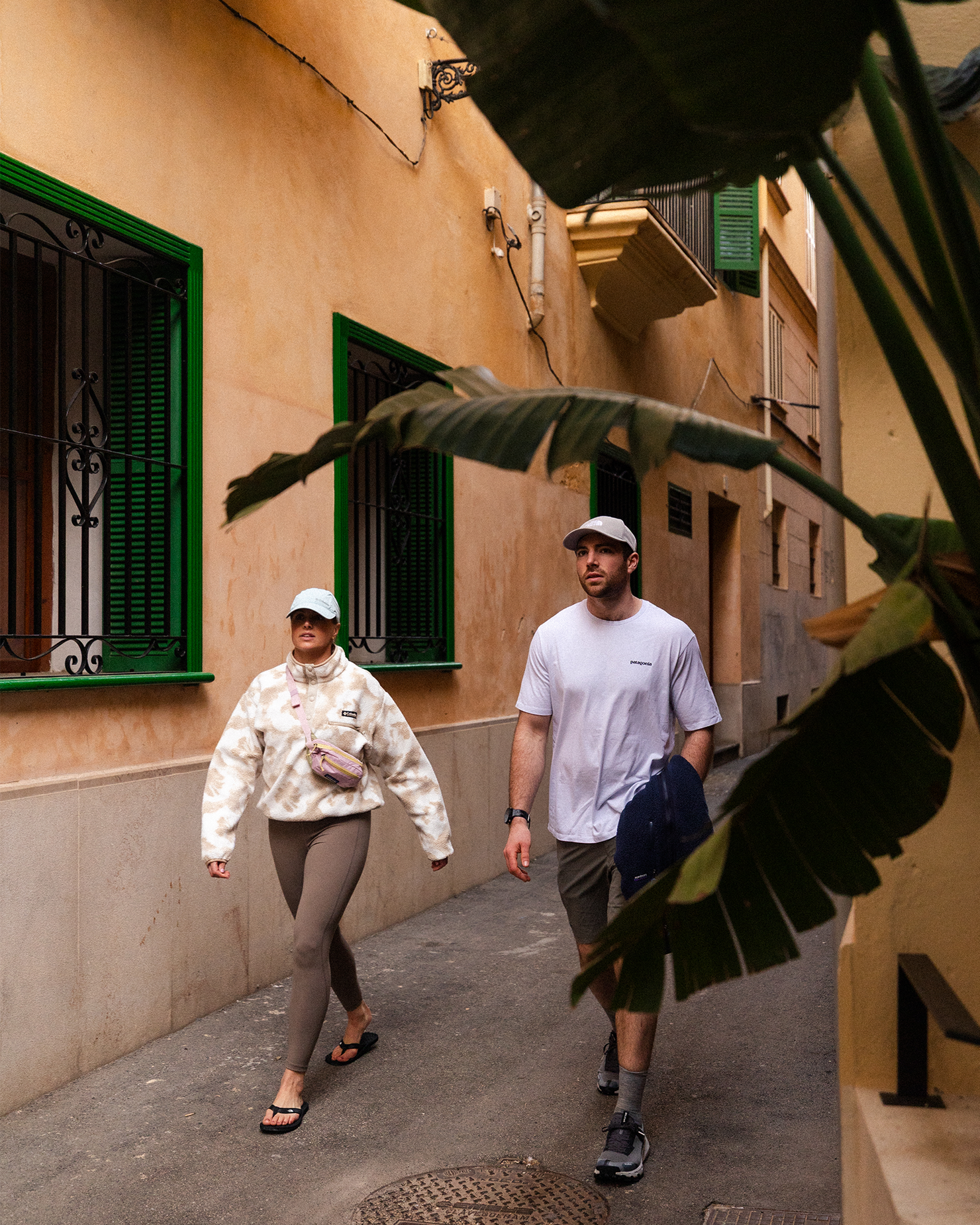 A man and woman in casual attire walk down a narrow, sunlit street lined with beige walls and green windows, partially obscured by tropical leaves.