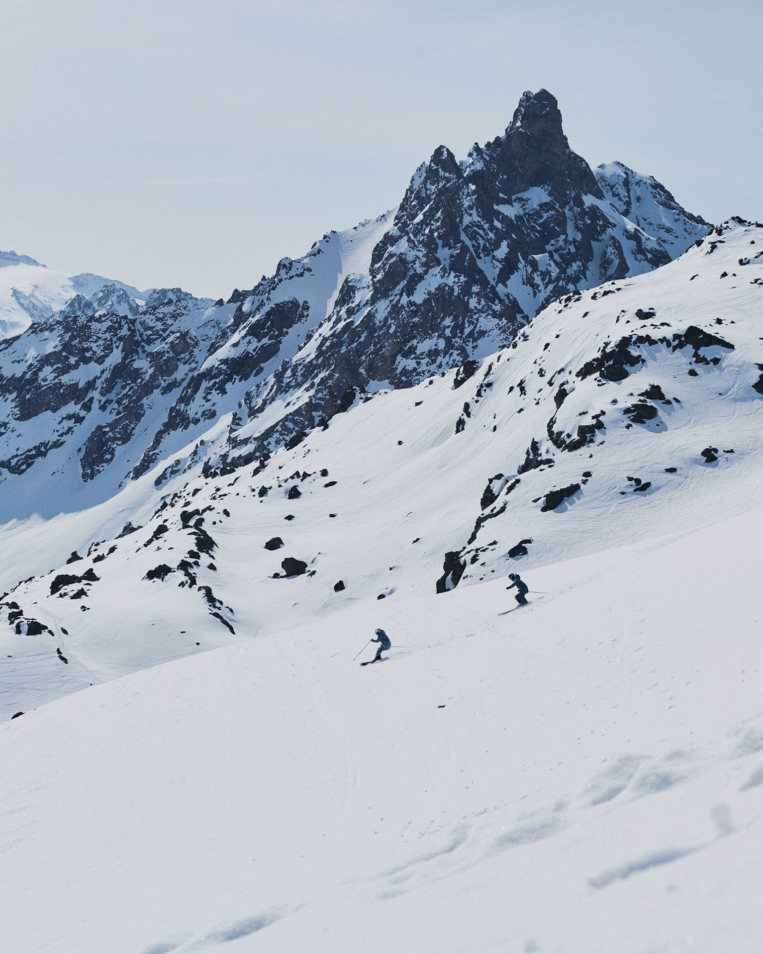 Two skiers in blue gear navigate down a snowy mountain slope, set against a backdrop of rugged, snow-covered peaks under a clear sky.