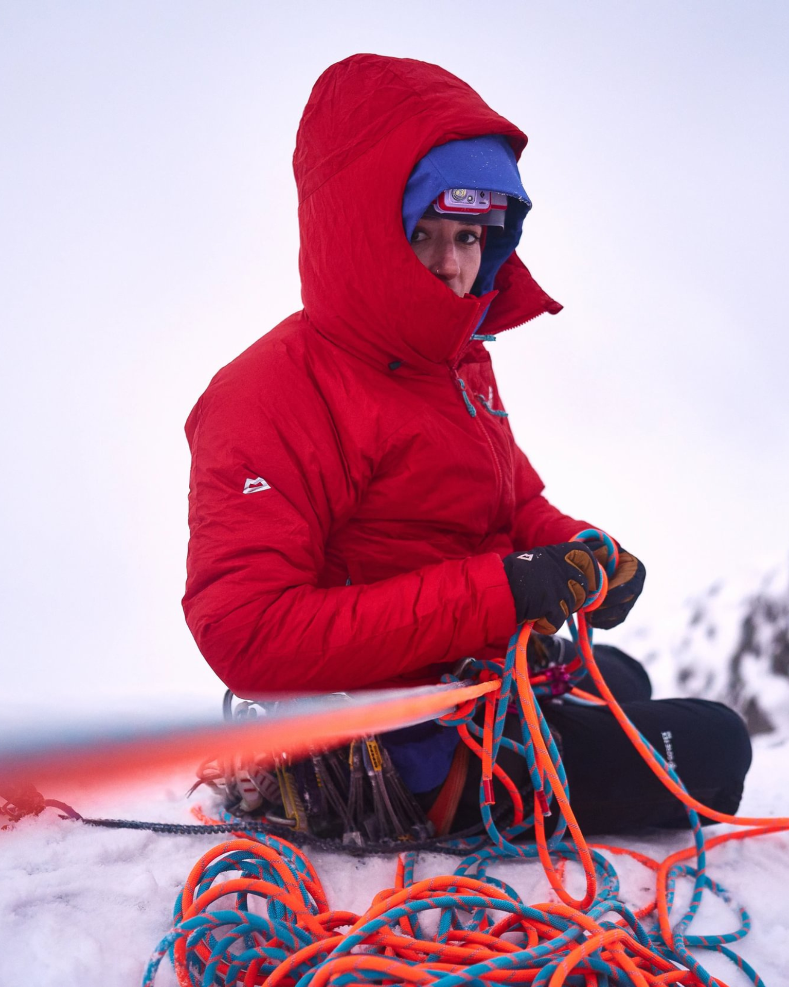 Female climber in red Mountain Equipment Alpamayo jacket belays on a snow-covered mountain slope, focused on a task.
