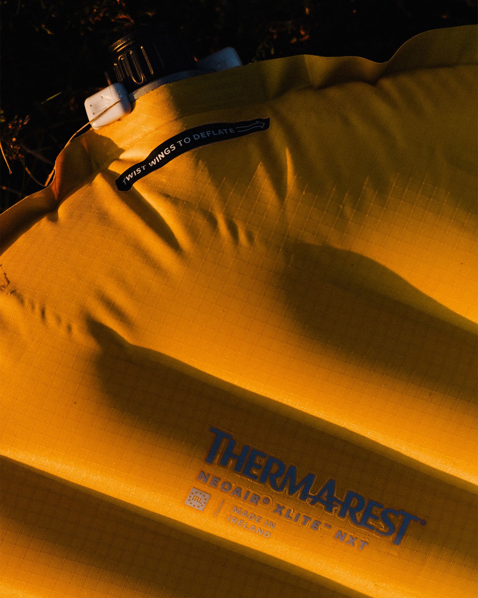 Close-up of a yellow Therm-a-Rest NeoAir Xlite NXT sleeping pad with a valve labeled "Twist Wings to Deflate," in warm lighting.