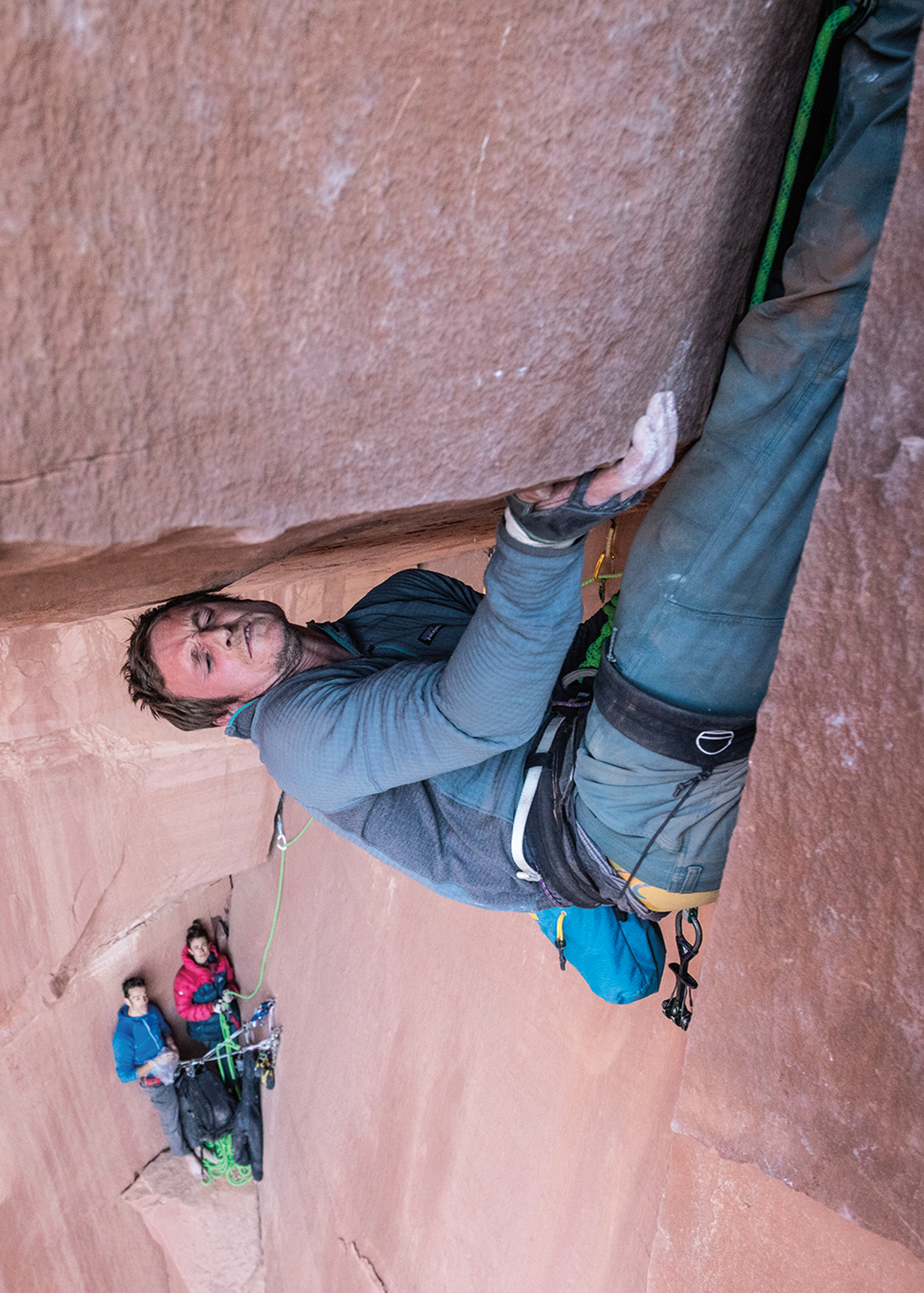 Climber in blue jacket ascends a vertical rock face; two people below secure the rope.